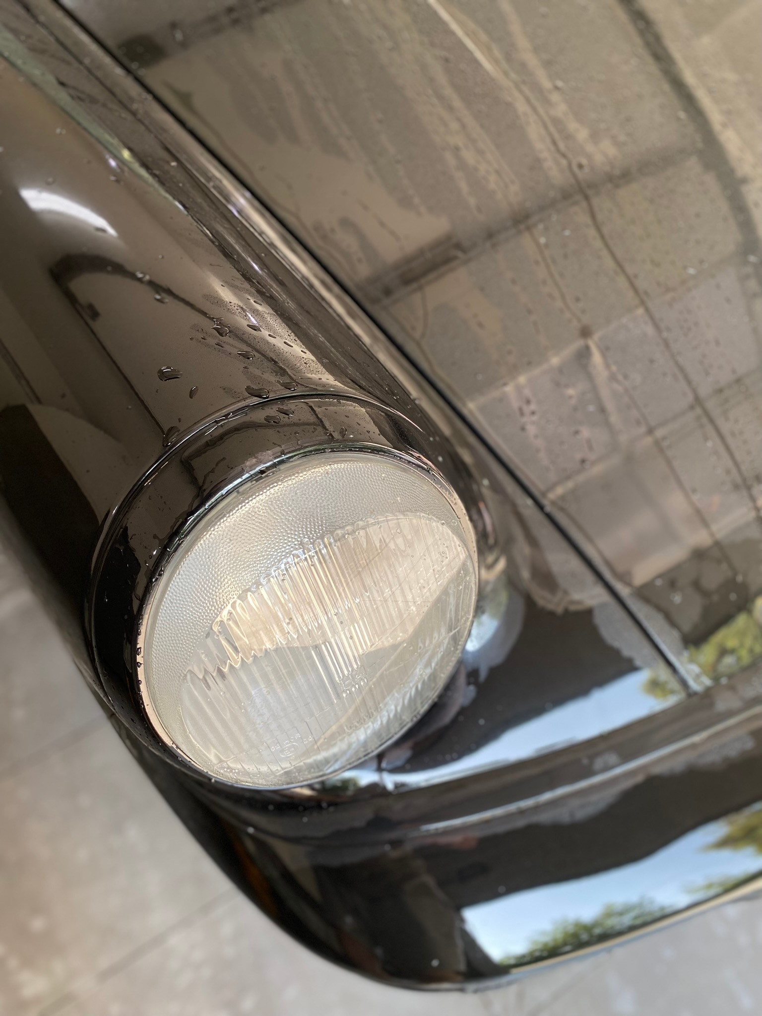 Reflektor klasycznego, czarnego samochodu z kroplami wody po myciu, widoczne odbicie drzew w lakierze.