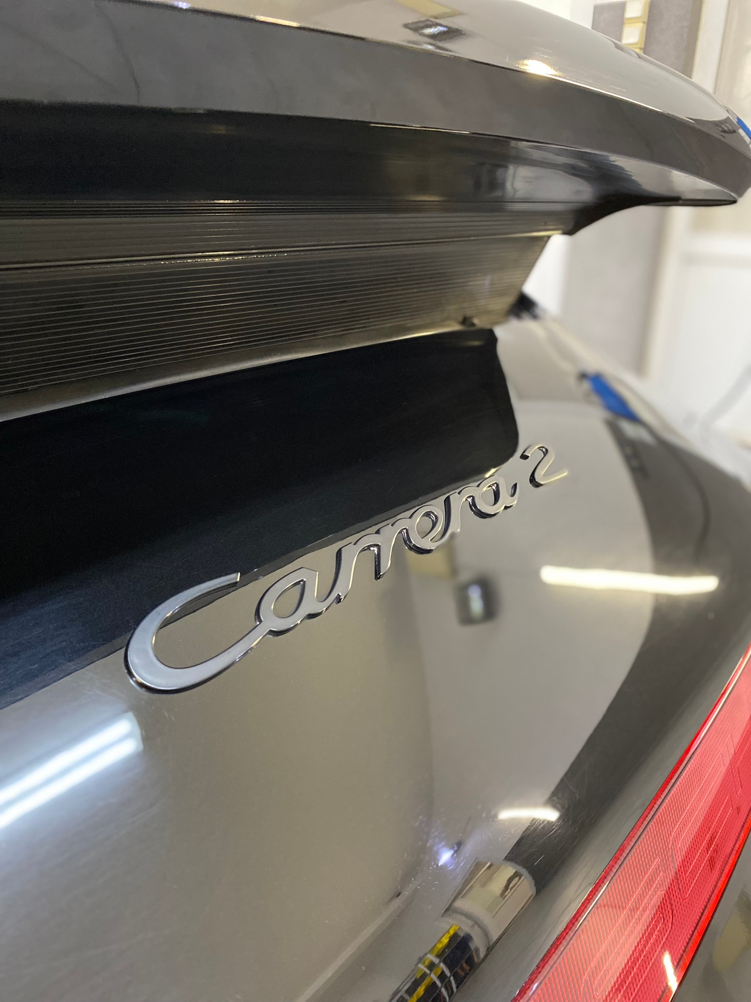 Błyszczący, czarny Porsche Carrera 2 z chromowanym napisem, widoczne odbicia światła na lakierze, fragment tylnej lampy i aplikator do polerowania lakieru.
