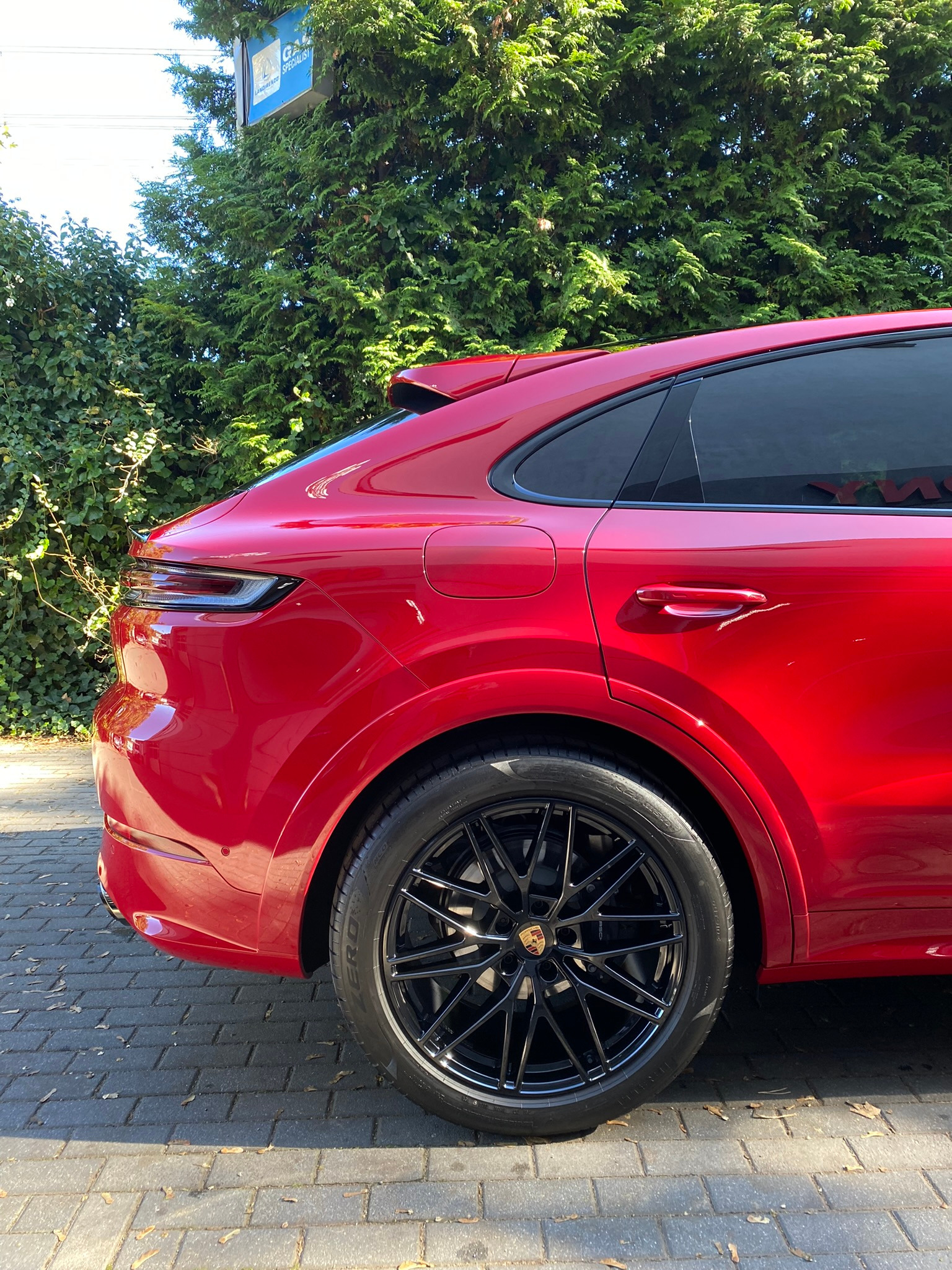 Czerwony samochód typu SUV, prawdopodobnie Porsche Cayenne, z czarnymi felgami, zaparkowany na brukowanej powierzchni w Gdańsku, z zielonymi krzewami w tle. Widoczny fragment karoserii po detailing.
