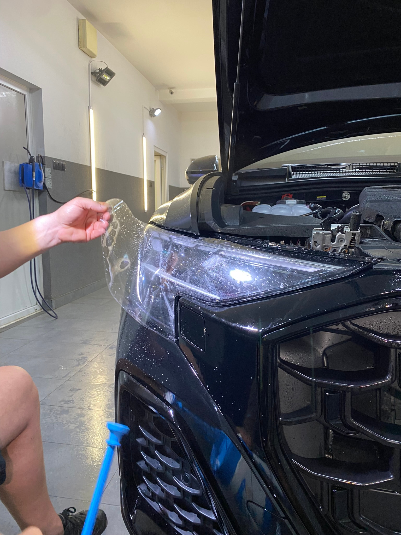 Nakładanie przezroczystej folii ochronnej na reflektor czarnego samochodu z otwartą maską, widoczne narzędzie do aplikacji i detale maskownicy