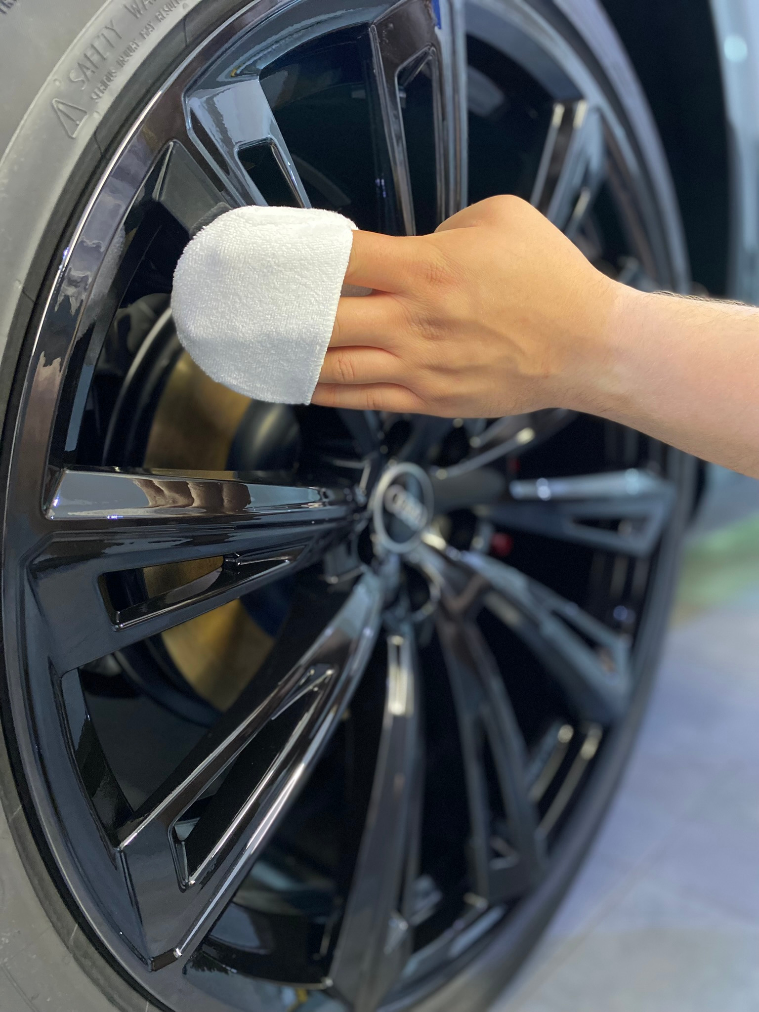 Czyszczenie czarnej felgi aluminiowej samochodu marki Audi białą ściereczką z mikrofibry, widoczne logo producenta, opona i tarcza hamulcowa.