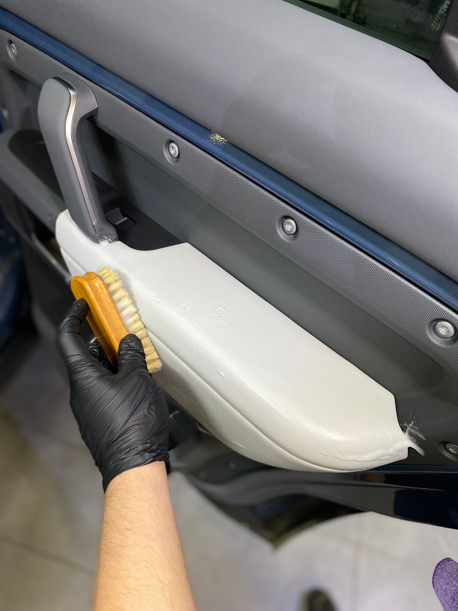 Szczotkowanie jasnej tapicerki drzwi samochodowych, widoczna piana czyszcząca i ręka w czarnej rękawicy ochronnej.
