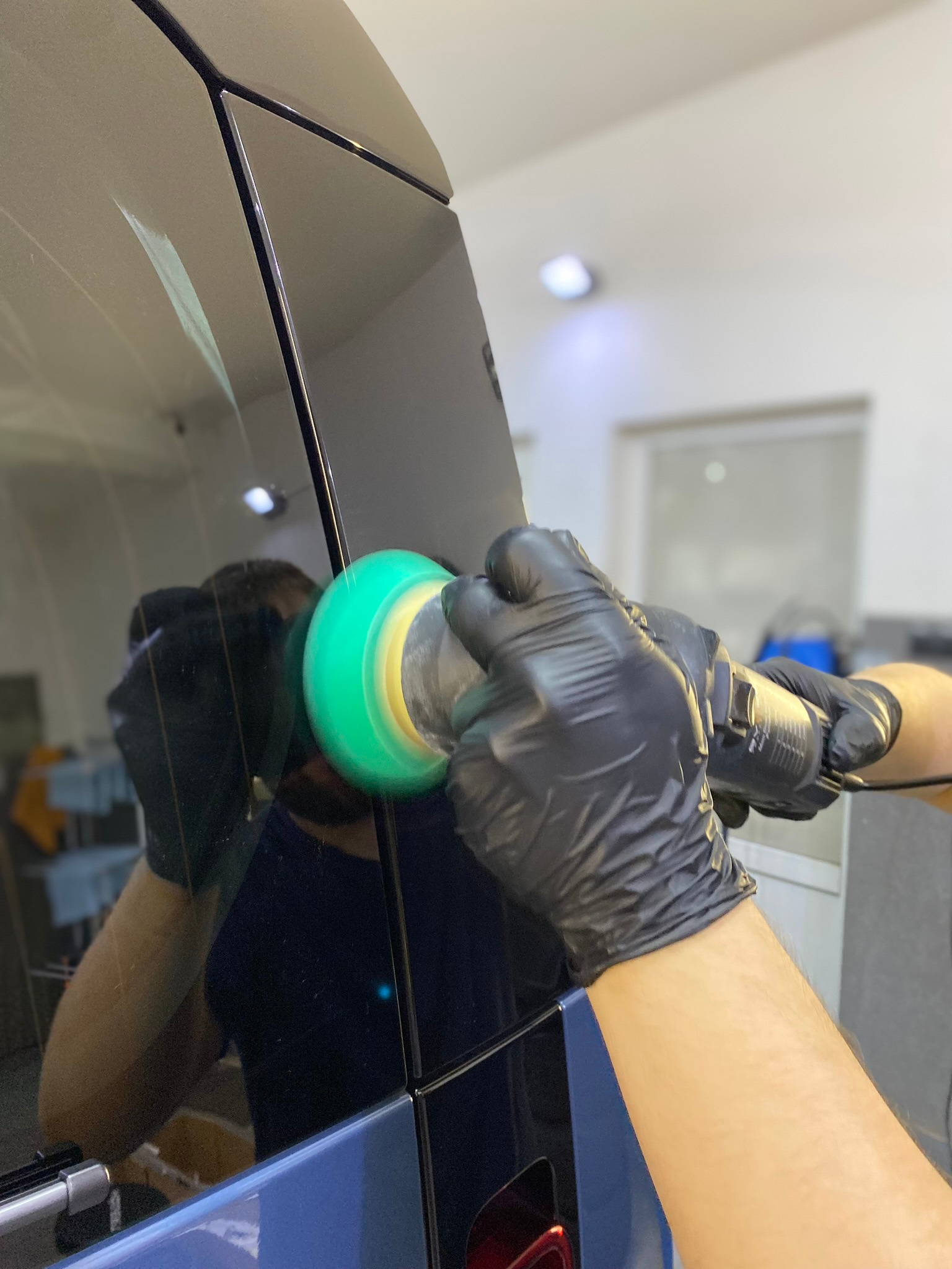 Polerowanie lakieru na czarnym samochodzie przy użyciu elektrycznej polerki z zieloną nakładką, praca wykonywana w rękawicach ochronnych.