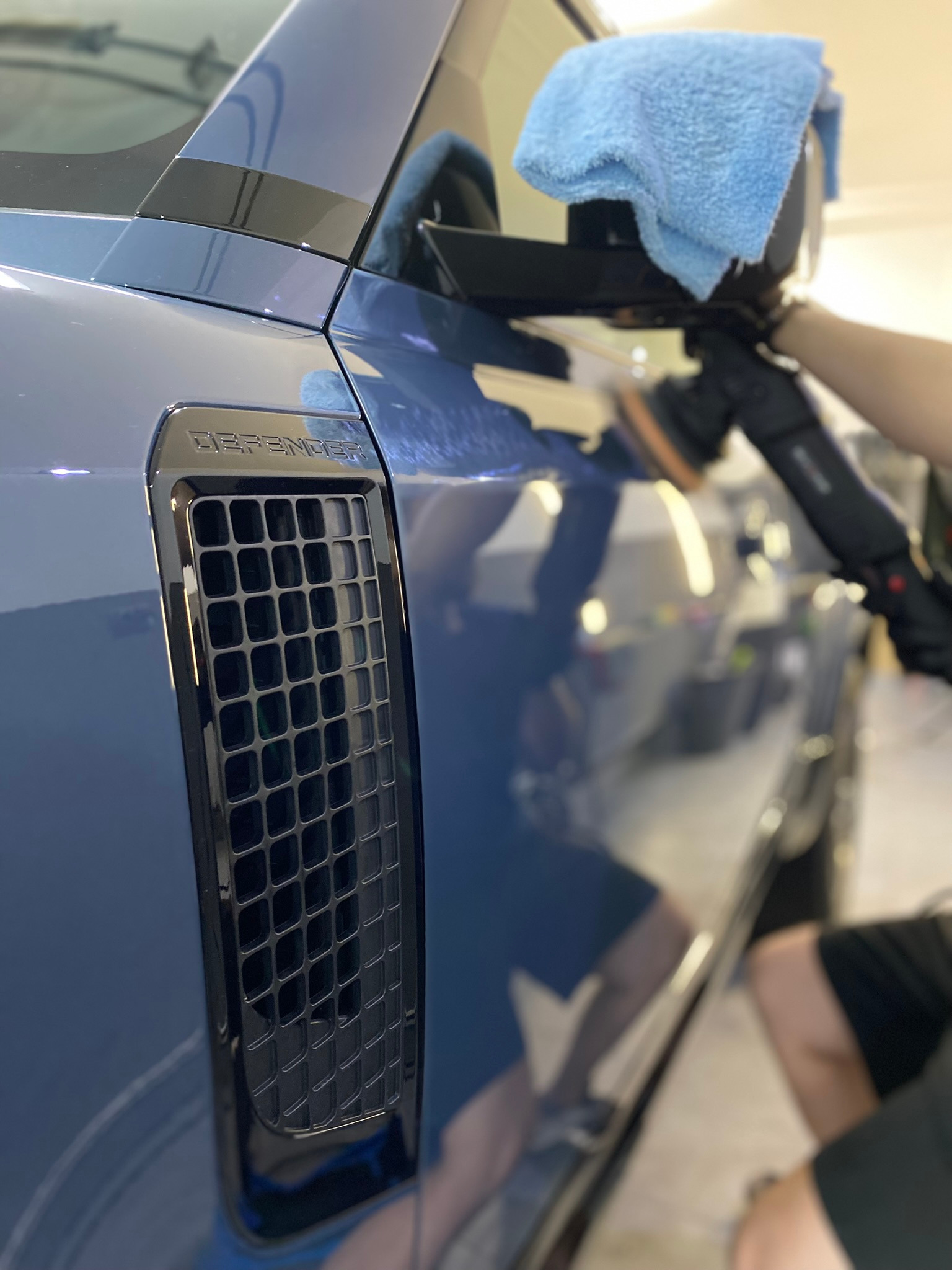 Błękitny Defender polerowany maszynowo przy użyciu niebieskiej mikrofibry, widoczny fragment karoserii z charakterystycznym grillem bocznym.