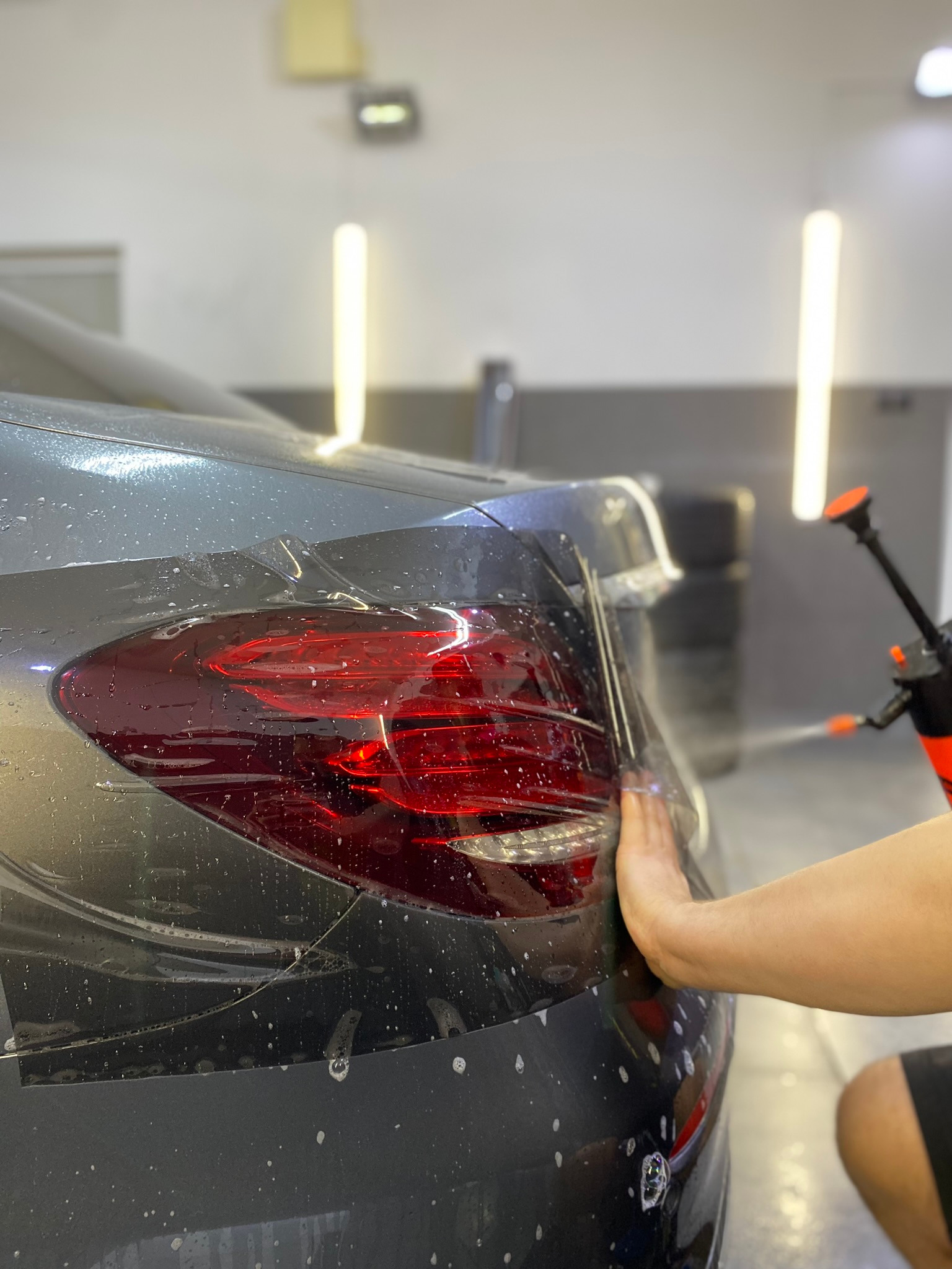 Aplikacja folii ochronnej na tylną lampę samochodu w jasnym warsztacie, widoczne krople wody i dłoń pracownika wygładzająca folię. W tle butla ze spryskiwaczem.