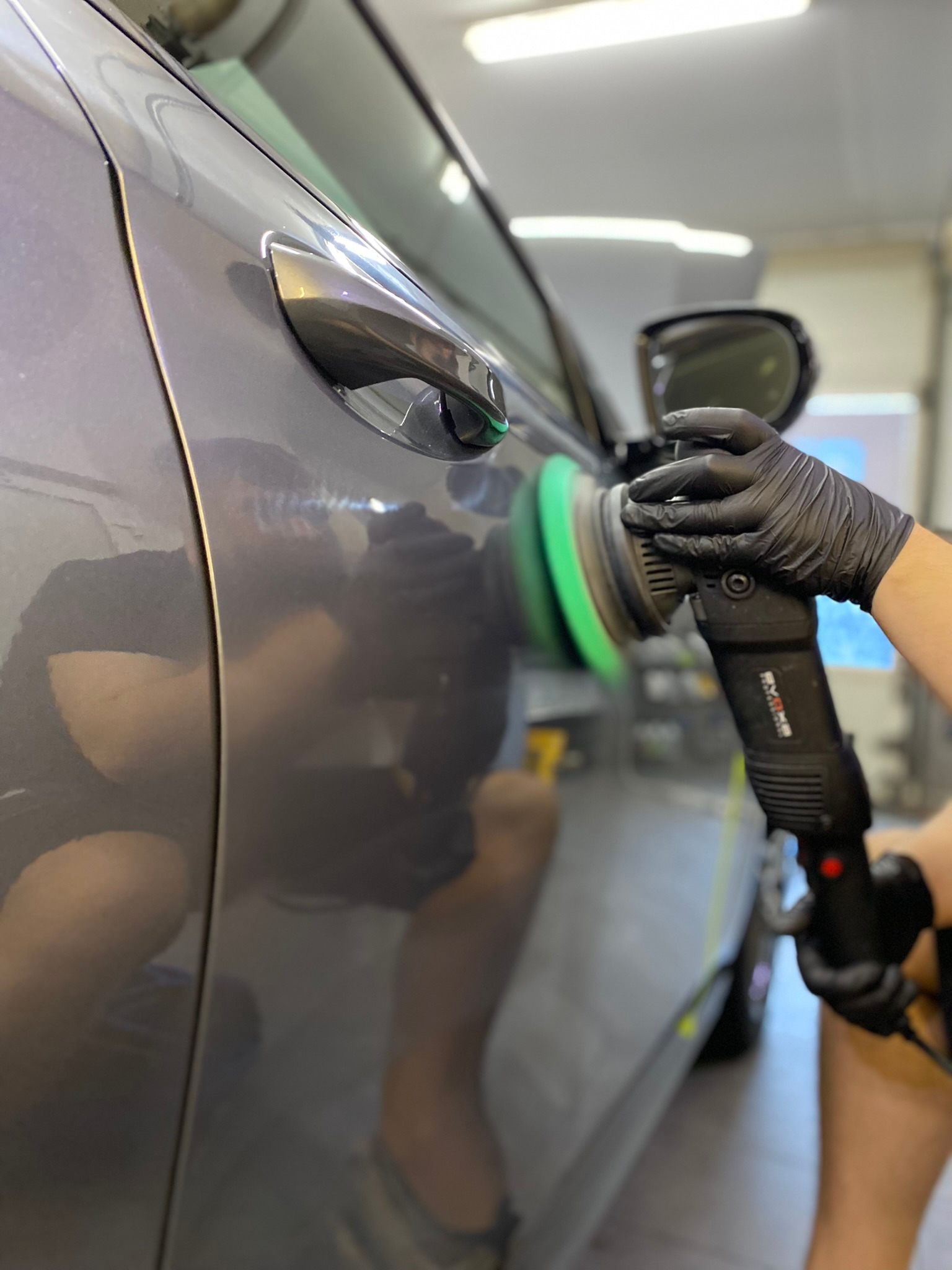Polerowanie boku szarego samochodu za pomocą polerki elektrycznej z zieloną nakładką, dłoń w czarnej rękawicy ochronnej, widoczne refleksy na lakierze.