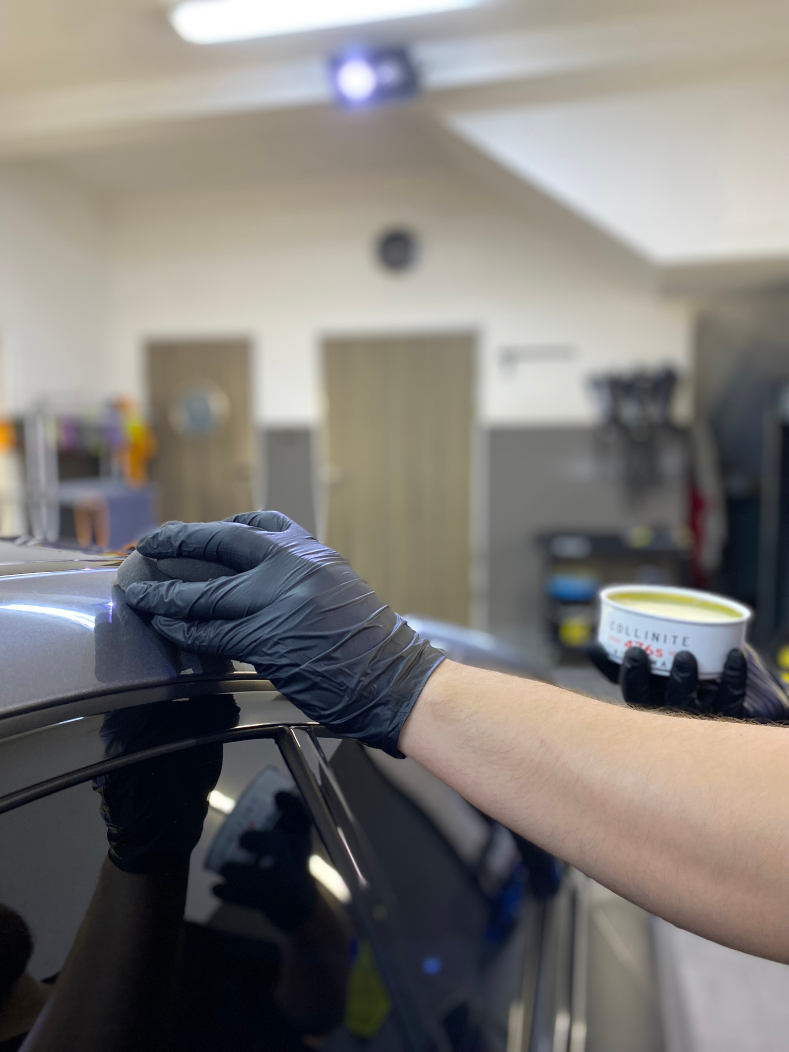 Nakładanie wosku ochronnego Collinite na lakier samochodu przez detailera w czarnych rękawiczkach, widoczny refleks światła na powierzchni.