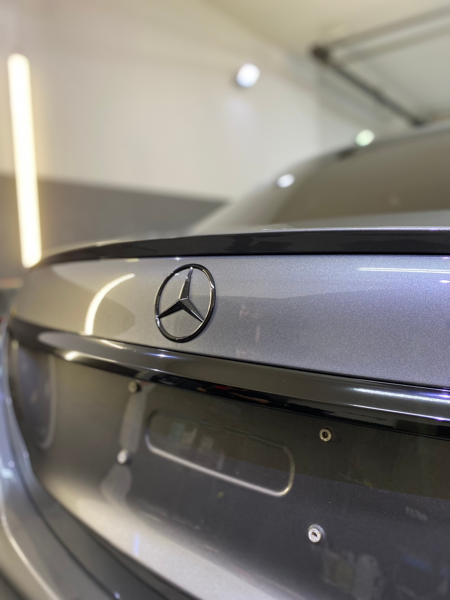 Szare auto marki Mercedes, widok na tylną klapę z czarnym spoilerem i logo marki, oświetlenie studyjne.