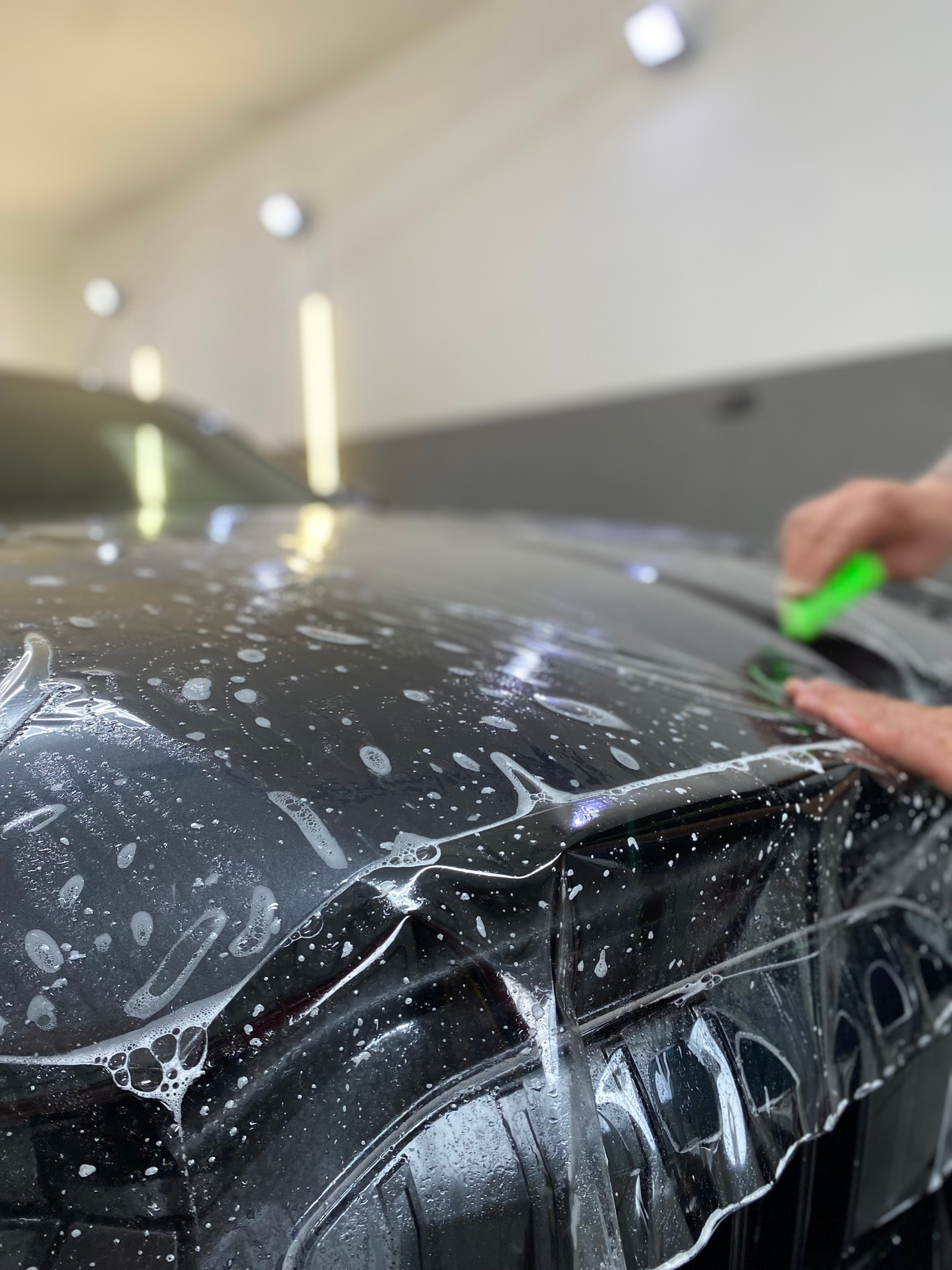 Aplikacja folii ochronnej na czarny samochód w warsztacie, widoczne bąbelki wody i narzędzie aplikacyjne w dłoni.