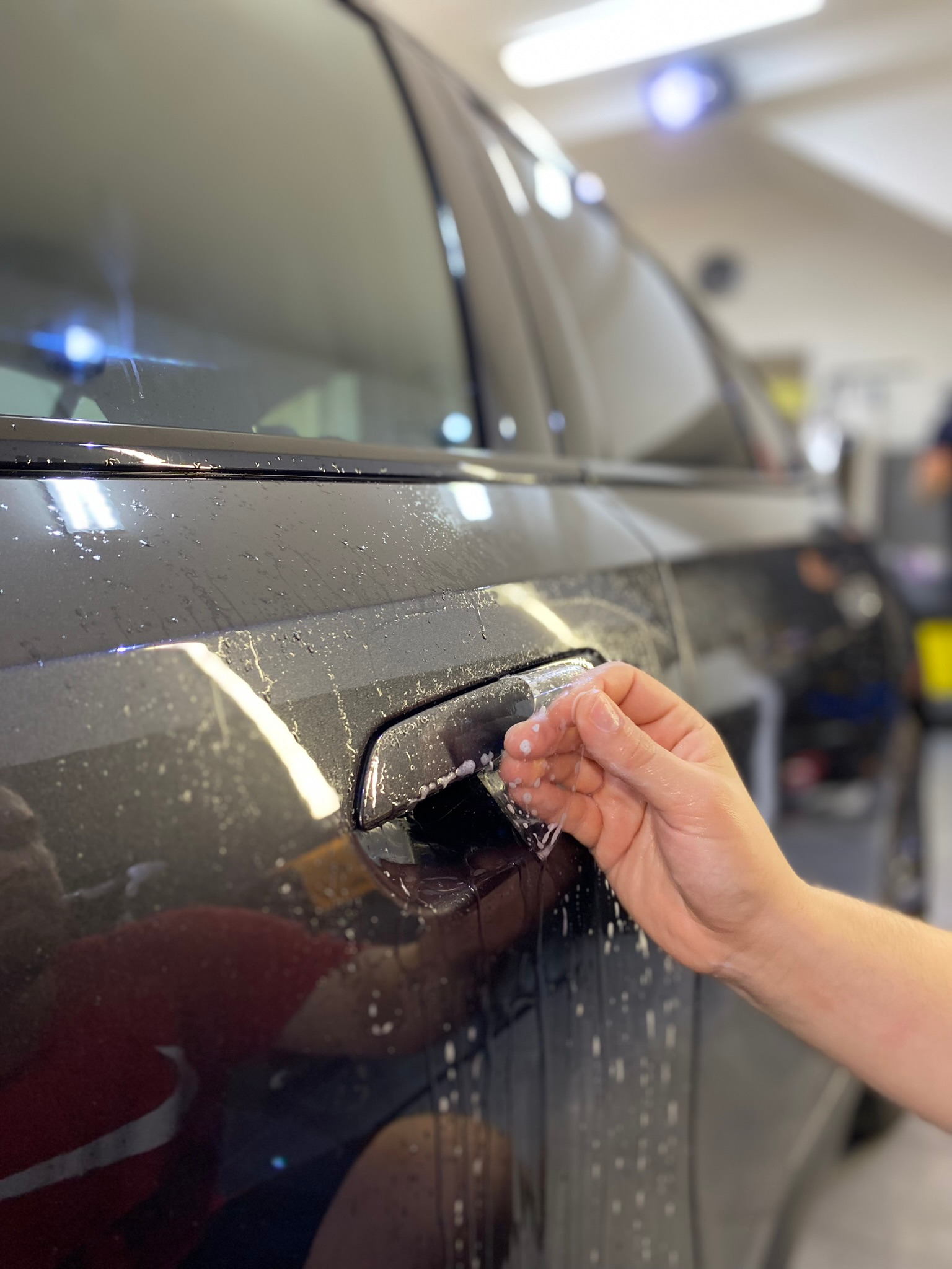 Aplikacja folii ochronnej na klamkę samochodu, widoczne krople wody i ręka z aplikatorem, w tle zamazane wnętrze warsztatu.