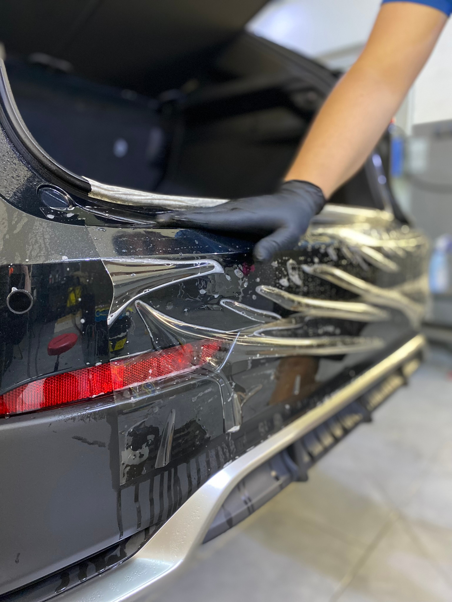 Nakładanie folii ochronnej na tylny zderzak czarnego samochodu, widoczna ręka w czarnej rękawicy wygładzająca folię, otwarty bagażnik, krople wody na powierzchni.