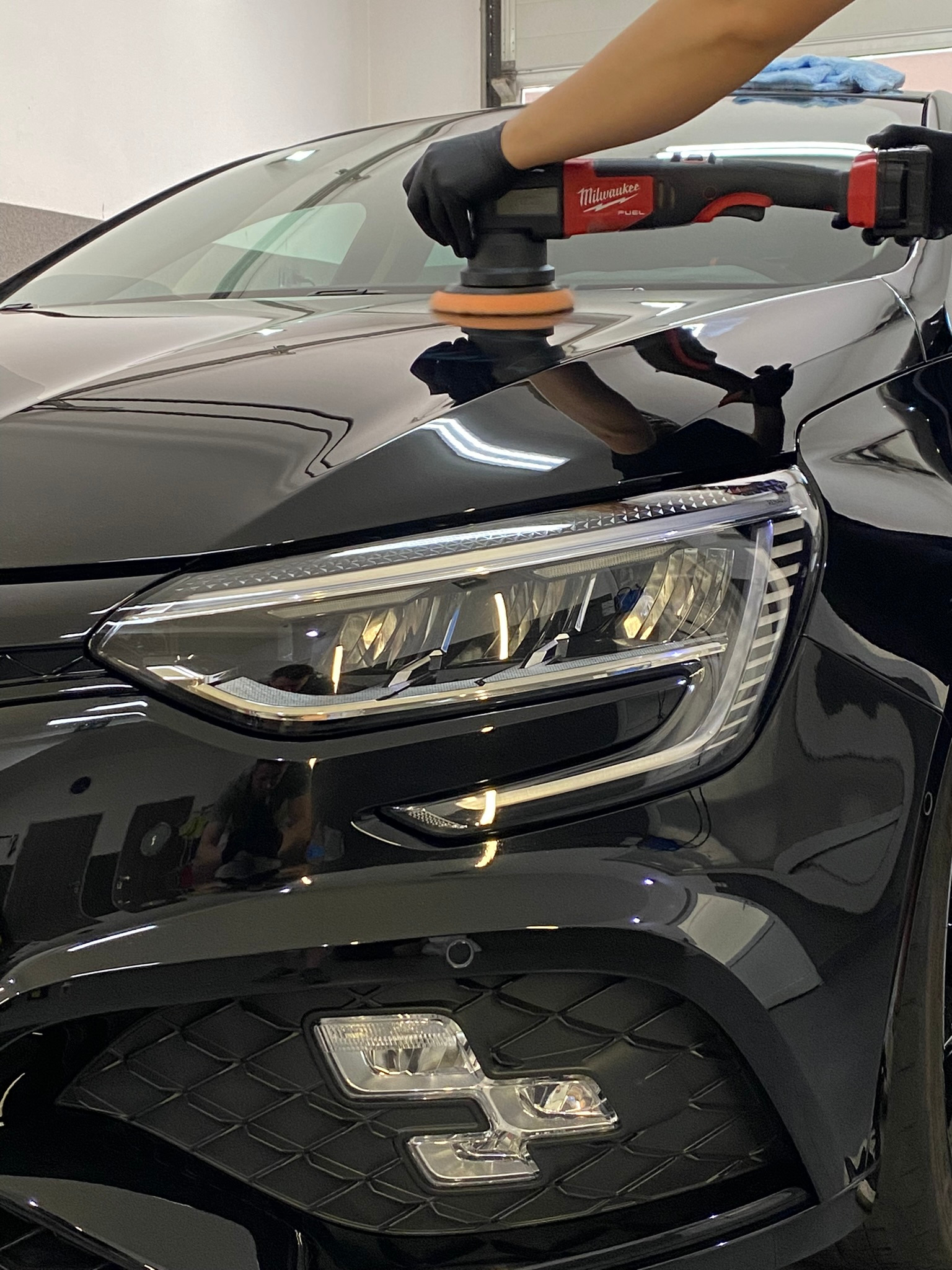 Polerowanie lakieru czarnego samochodu za pomocą polerki Milwaukee Fuel, widoczny reflektor i fragment maski, odbicie osoby w lakierze.