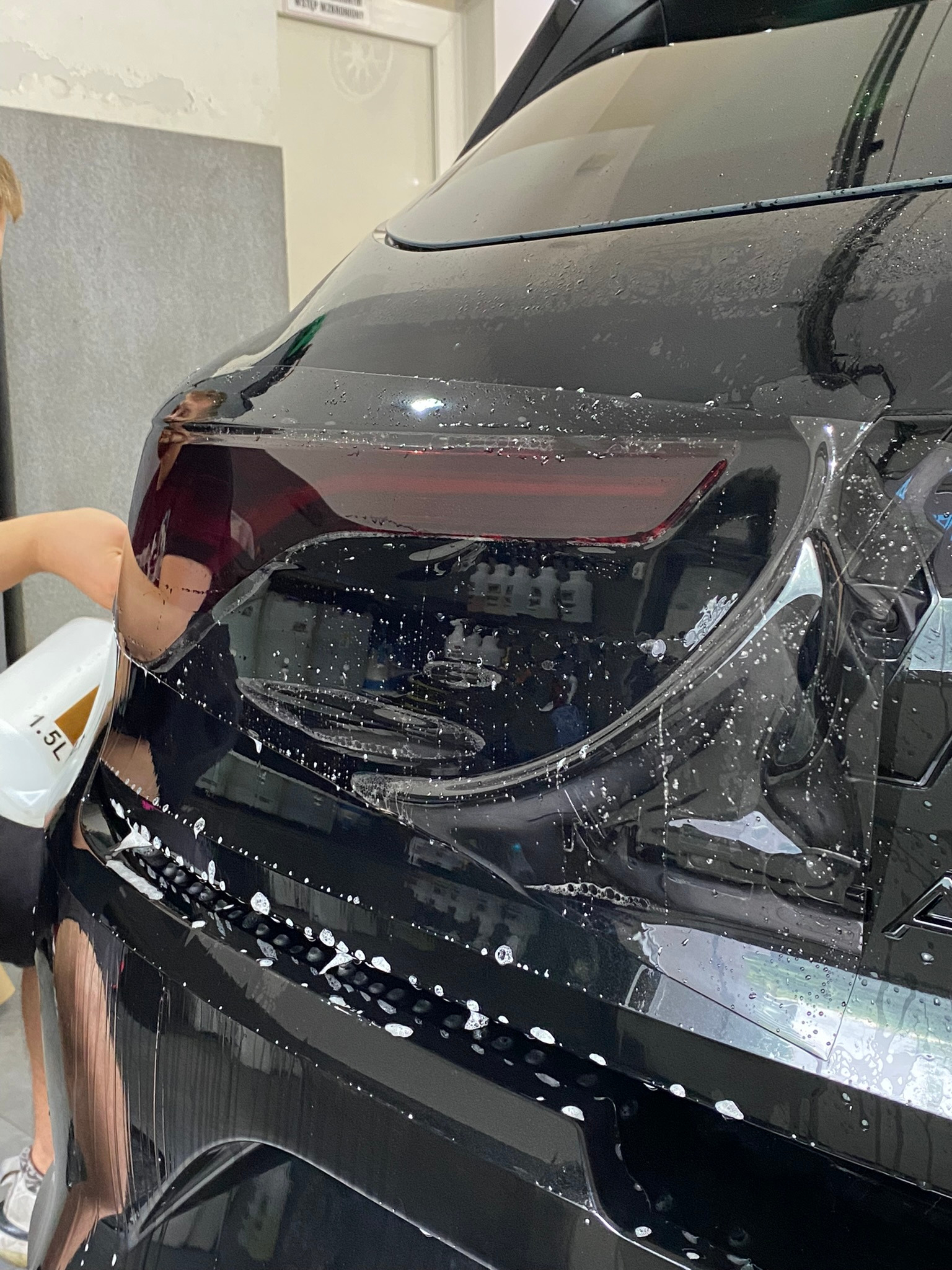 Aplikacja folii ochronnej na tylną lampę czarnego samochodu, widoczny pracownik z butelką 1.5L z roztworem, liczne pęcherzyki powietrza pod folią.