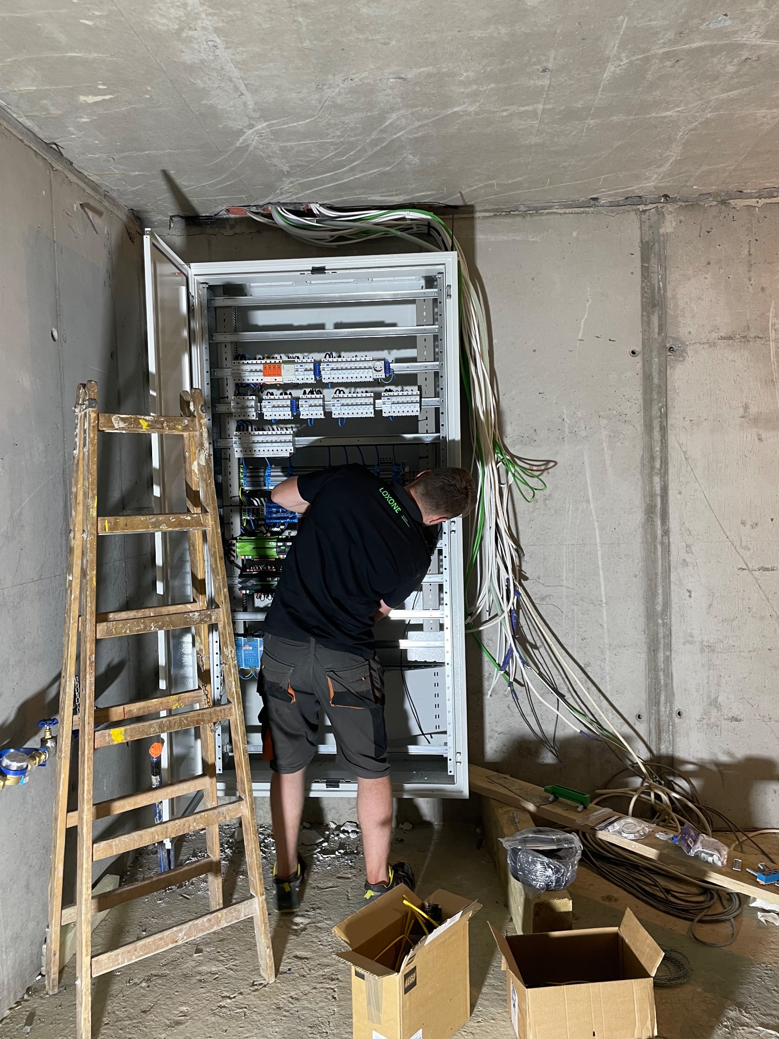 Elektryk w czarnej koszulce pochylony nad otwartą szafą rozdzielczą z przewodami w surowym wnętrzu, drabina oparta o ścianę, widoczne kable i skrzynki kartonowe na podłodze.