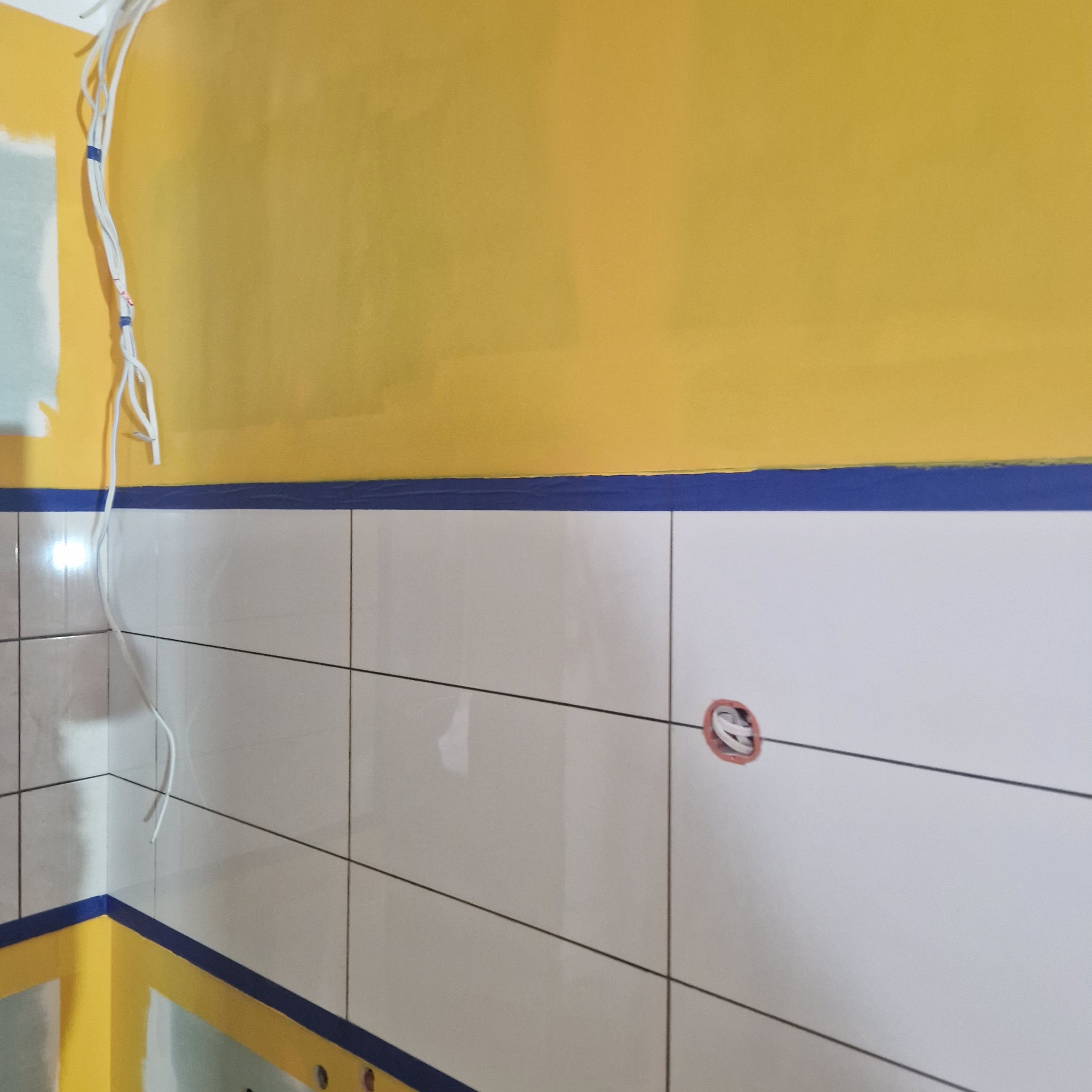 Żółta ściana w trakcie malowania z niebieską taśmą malarską nad białymi płytkami, widoczne kable elektryczne.