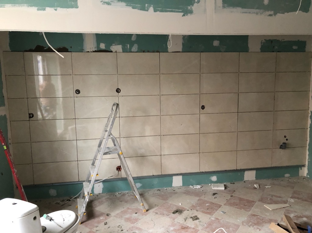 Ściana w trakcie układania beżowych, prostokątnych płytek ceramicznych w kuchni, widoczna drabina, poziomica i instalacja elektryczna.