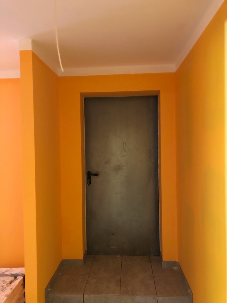 Ściany świeżo pomalowane na intensywny pomarańczowy kolor, szara metalowa futryna i drzwi, płytki podłogowe na schodku wejściowym, widoczny kabel zwisający z sufitu.