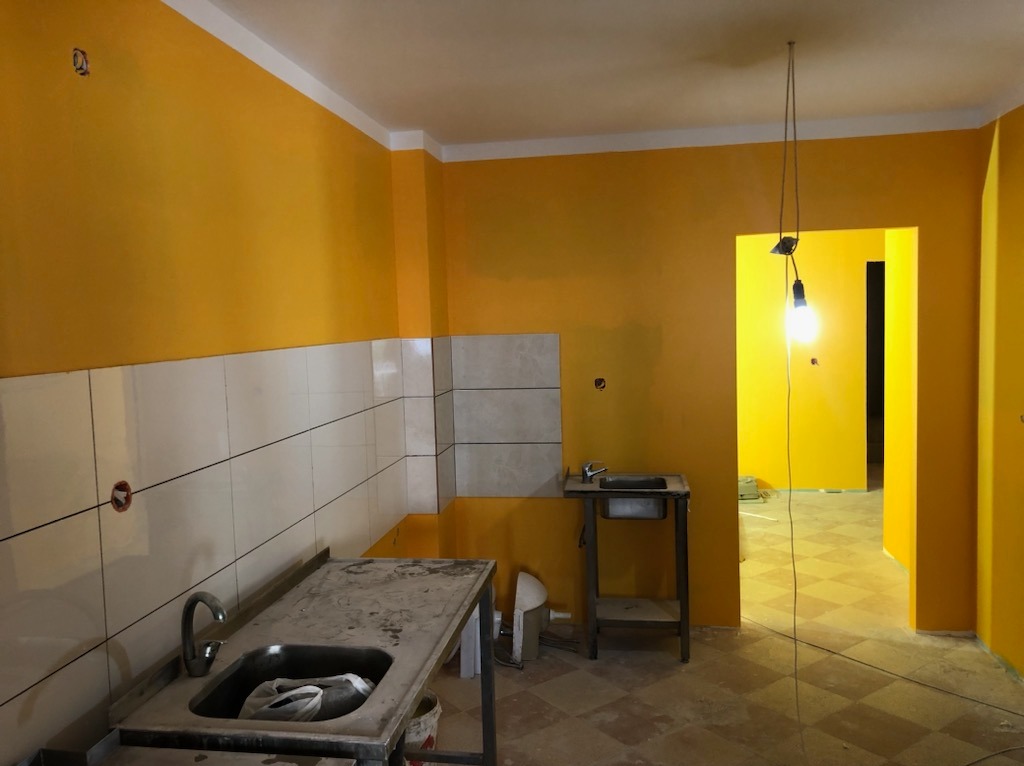 Żółta kuchnia w trakcie remontu: świeżo pomalowane ściany, częściowo ułożone płytki, prowizoryczne oświetlenie z żarówką na kablu.