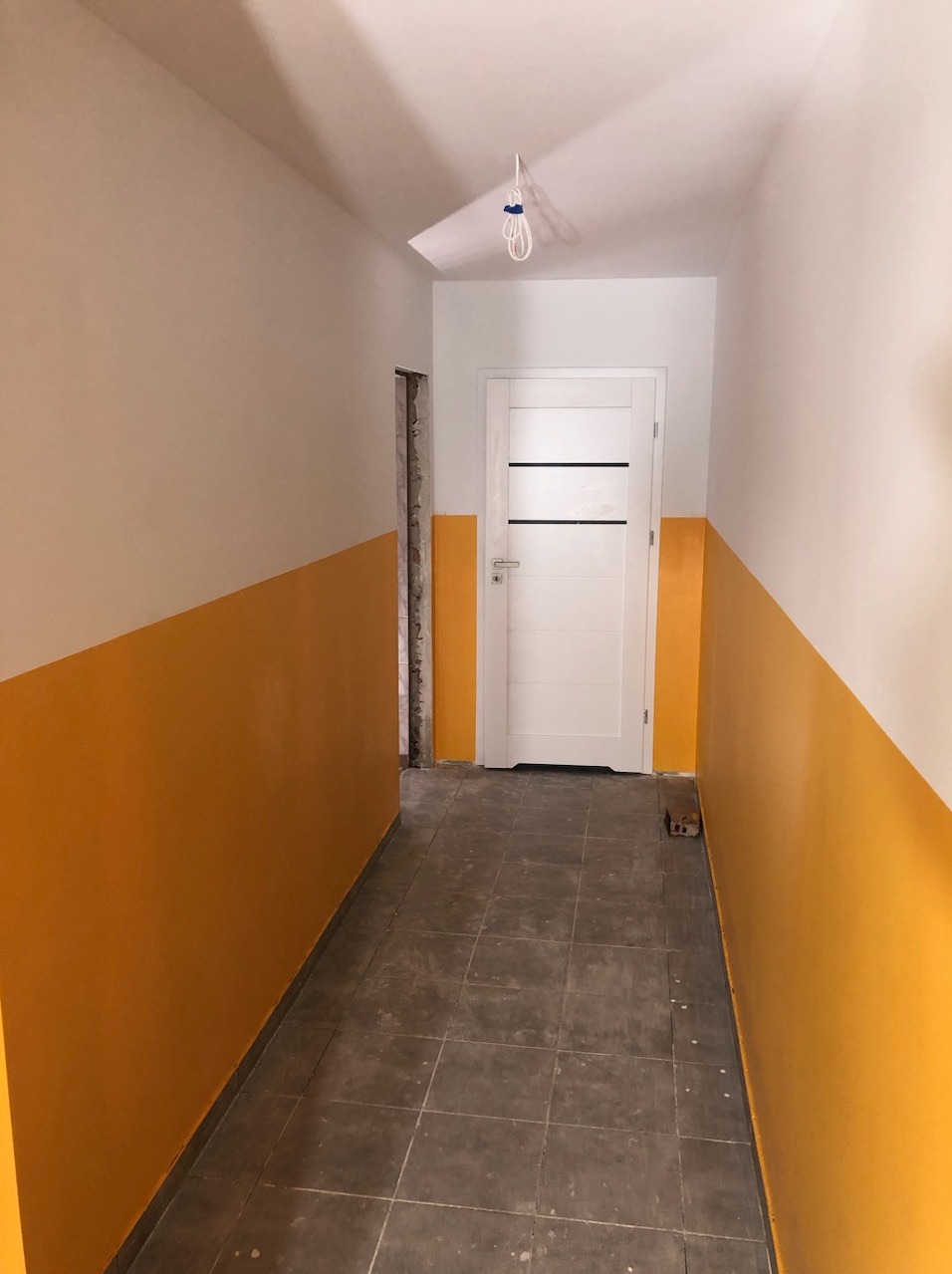 Korytarz po częściowym remoncie: pomalowane na żółto dolne partie ścian, białe górne partie i sufit, szare płytki na podłodze, białe drzwi z czarnymi paskami, wystające kable z sufitu.