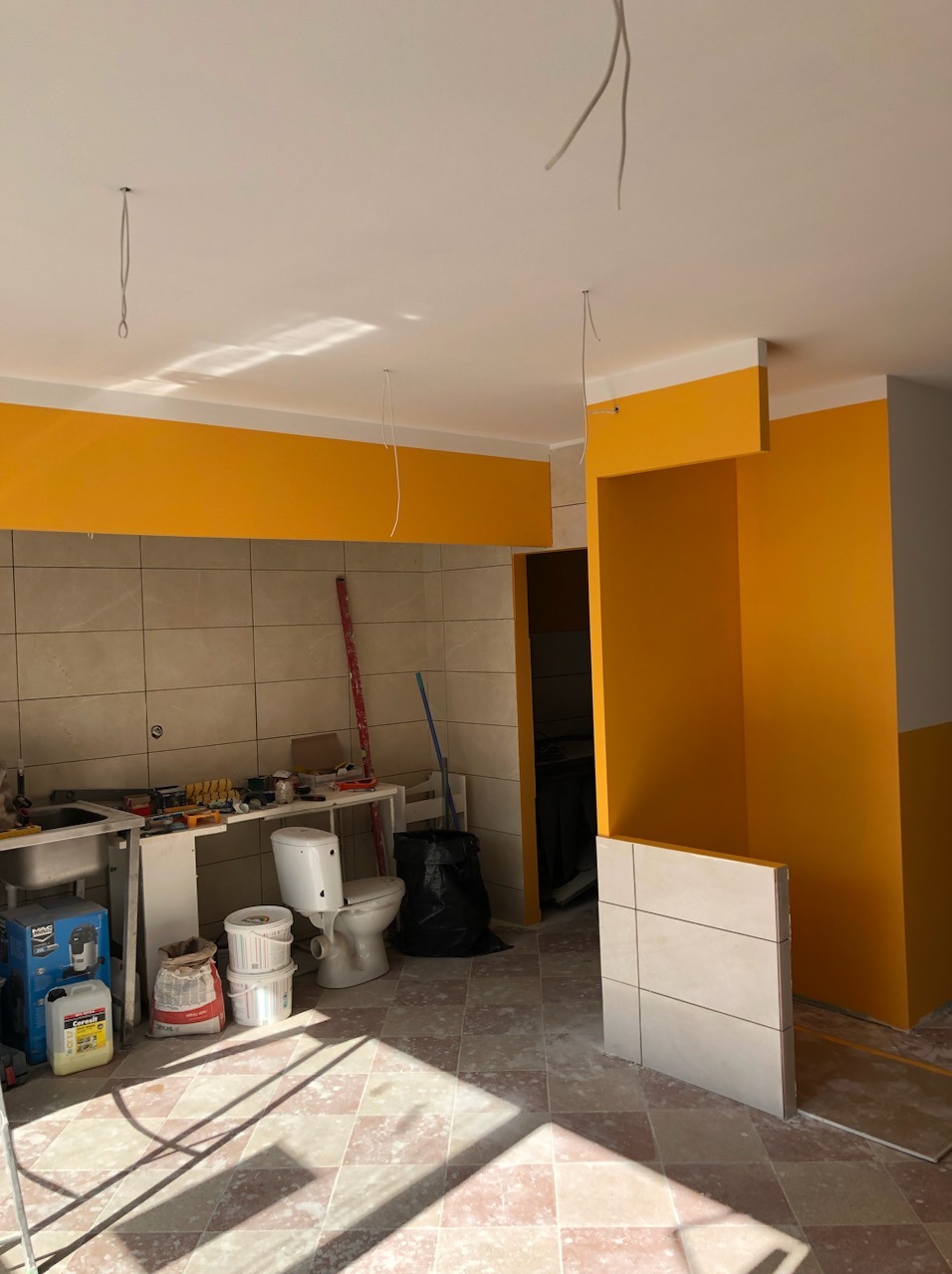 Remont łazienki w trakcie: żółte ściany, częściowo ułożone płytki, niepodłączona toaleta i narzędzia na stole roboczym.