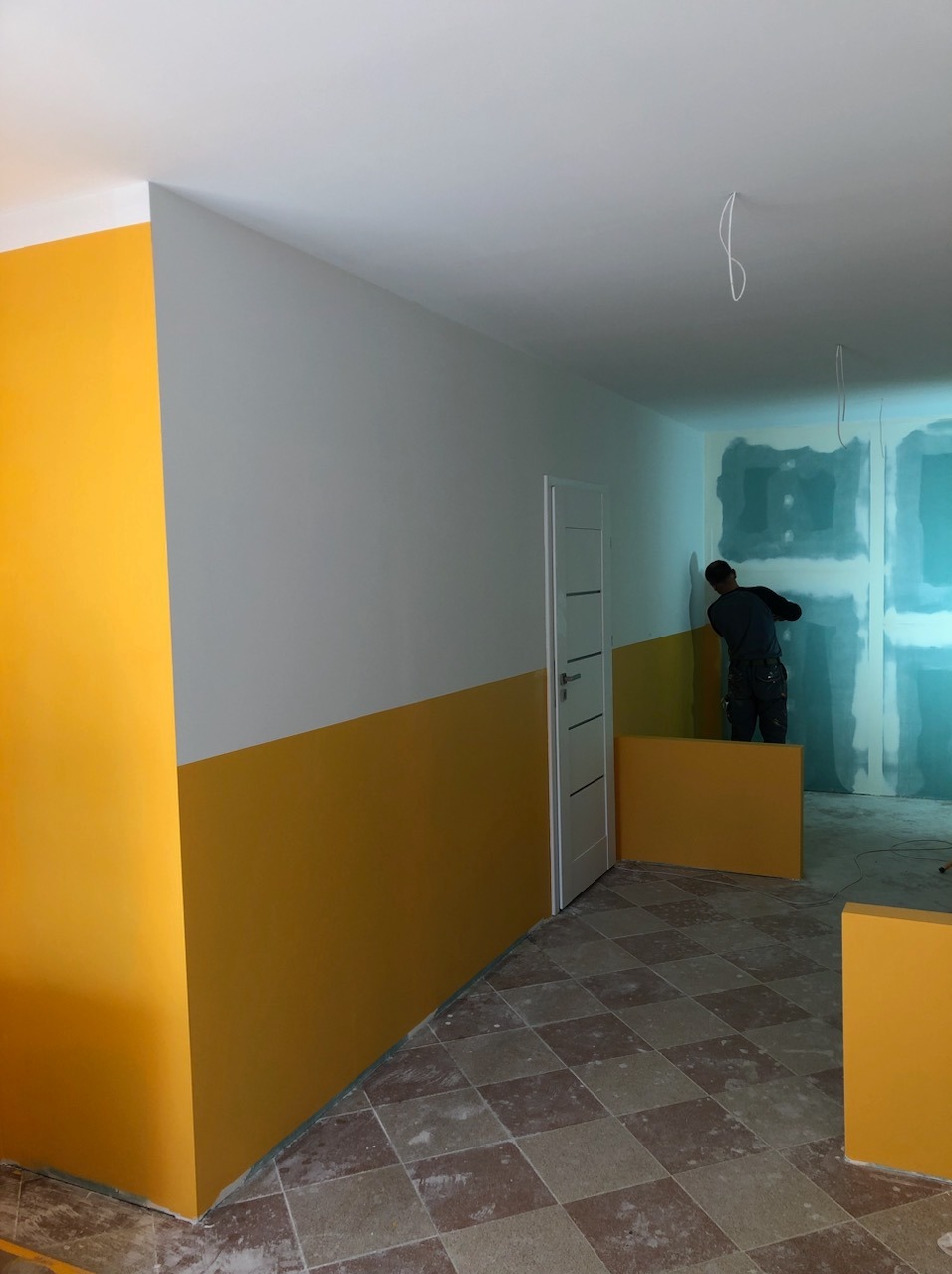 Dwukolorowe ściany w pomieszczeniu z płytkami podłogowymi w szachownicę; dolna część ścian pomalowana na żółto, górna na biało; w tle pracownik przygotowuje ścianę do malowania.