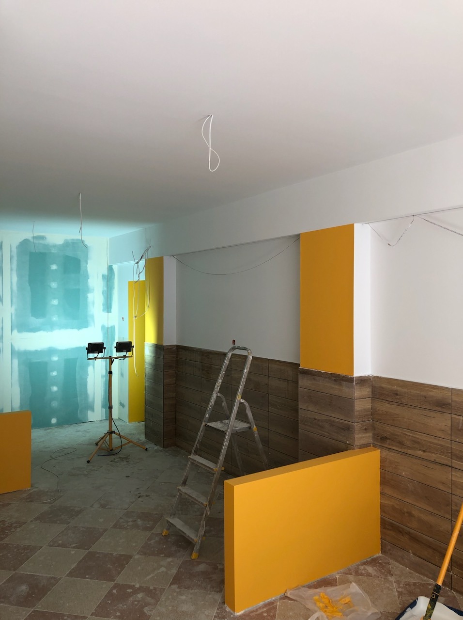 Wnętrze w trakcie prac remontowych: ściany częściowo pomalowane na żółto, fragmenty ścian pokryte płytami gipsowo-kartonowymi, widoczna drabina aluminiowa i lampa halogenowa.
