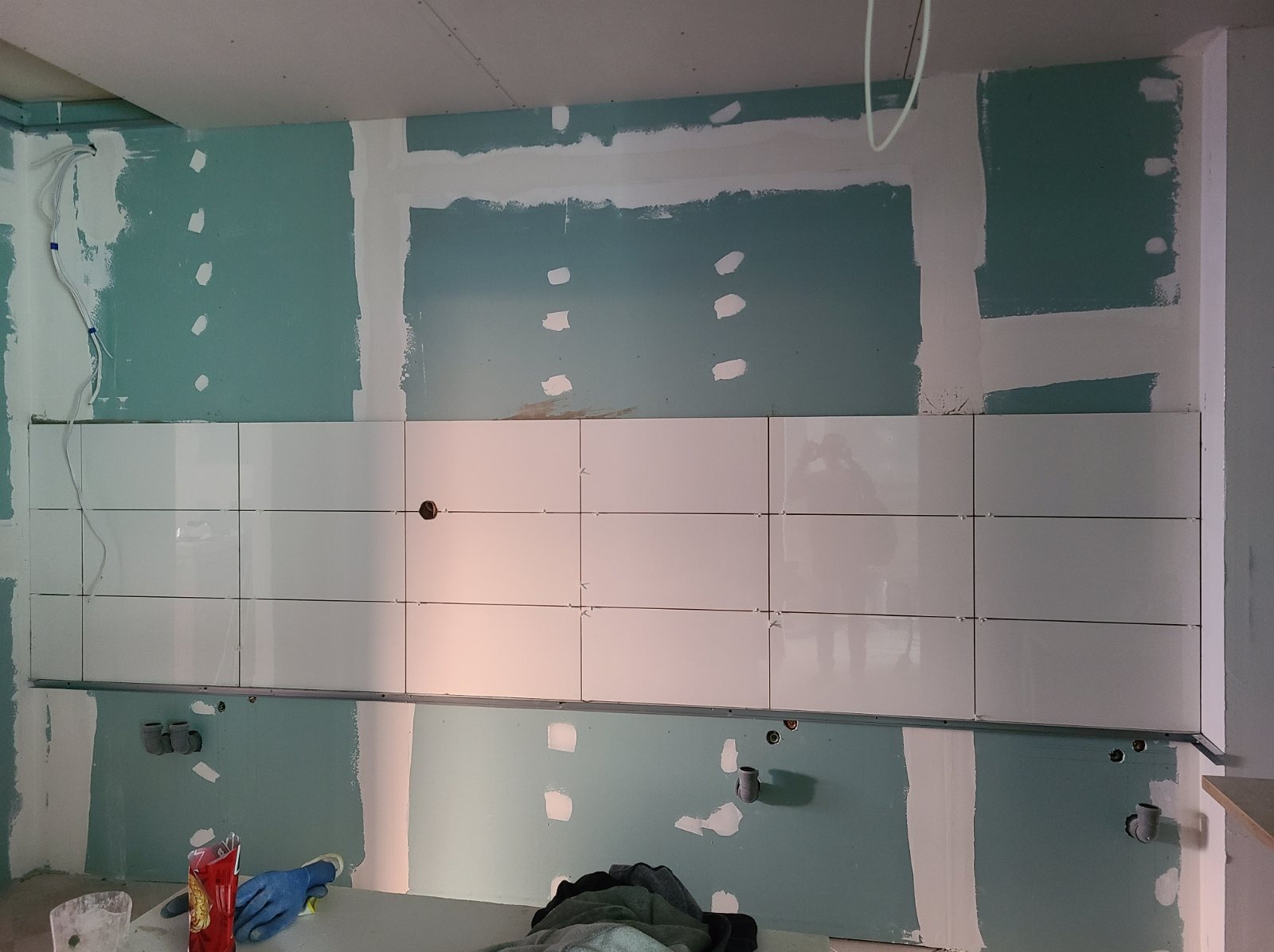 Ściana w trakcie remontu kuchni z częściowo położonymi białymi, błyszczącymi płytkami, widoczne profile montażowe, niezagruntowana płyta gipsowo-kartonowa i akcesoria robocze na blacie.