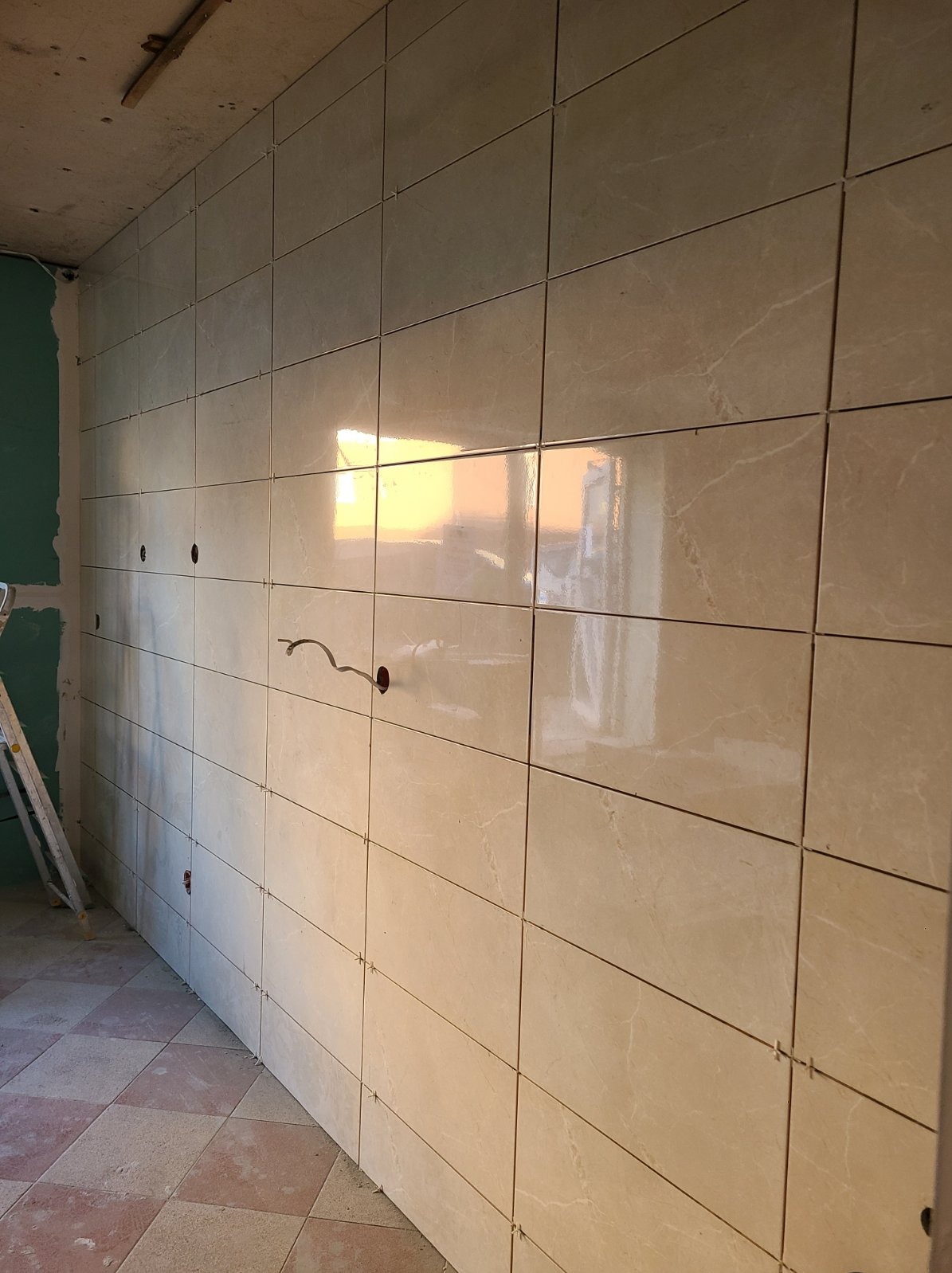 Ściana w trakcie układania płytek w kuchni lub łazience, widoczne otwory na instalacje elektryczne i krzyżyki dystansowe, podłoga częściowo wyłożona płytkami.