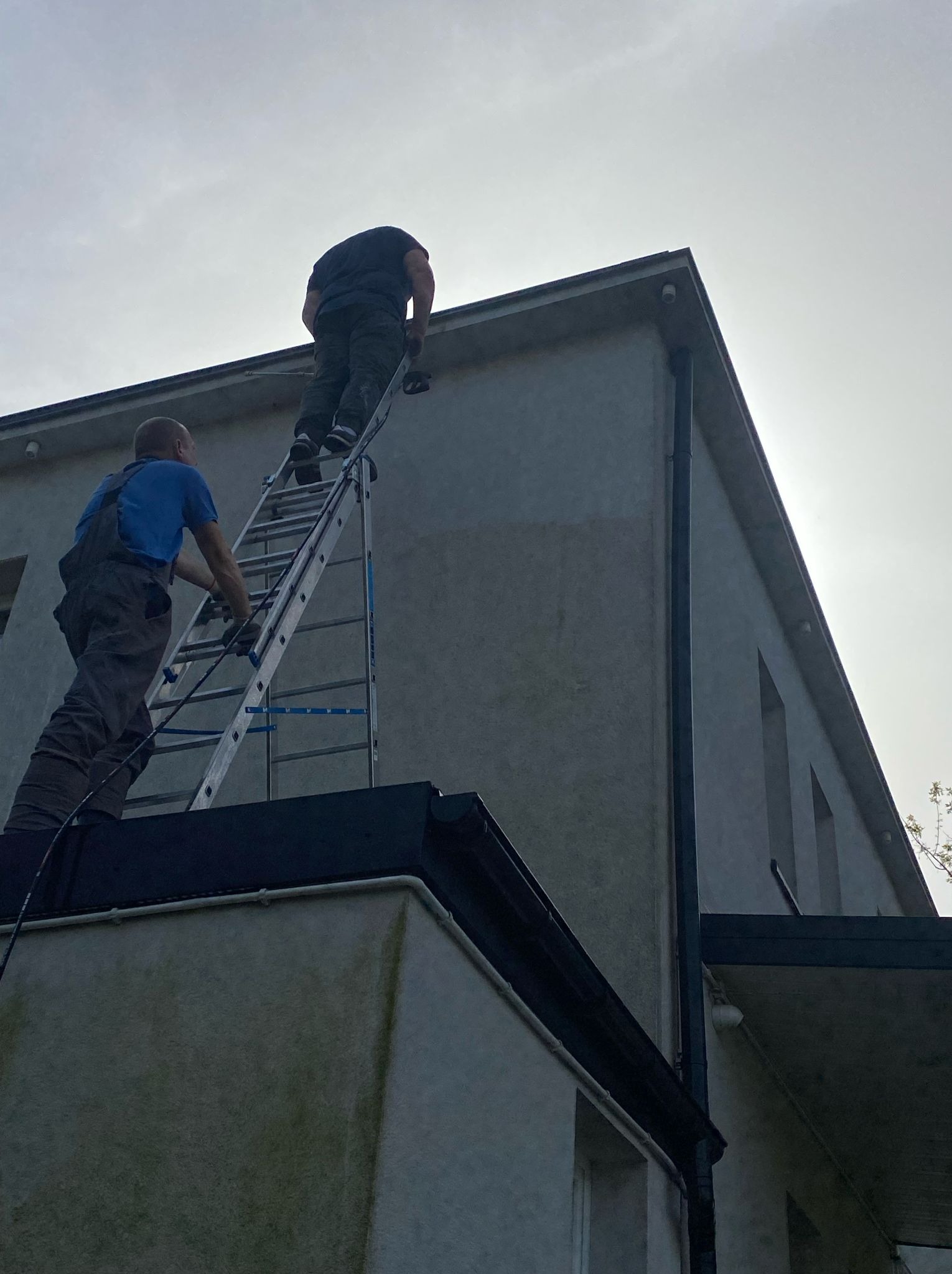 Dwóch mężczyzn na drabinie maluje elewację budynku z widocznym systemem rynnowym i oświetleniem zewnętrznym