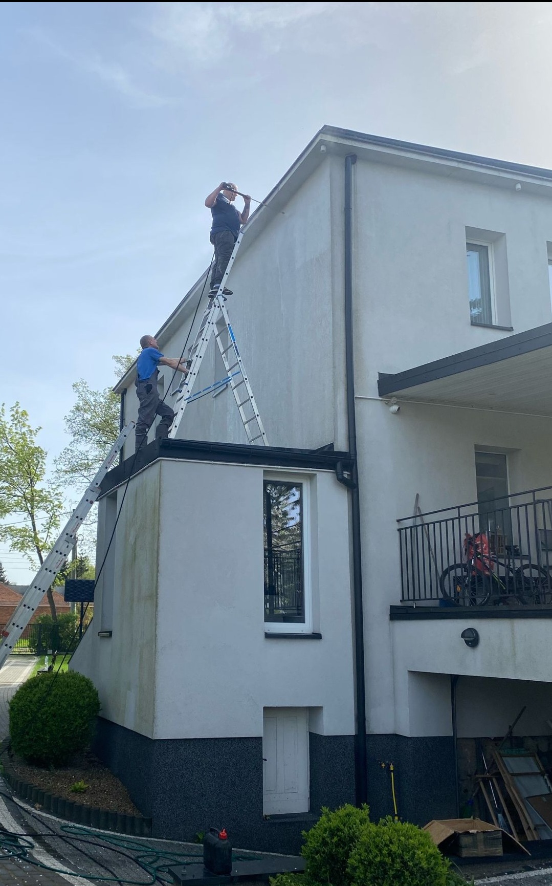 Dwaj mężczyźni na drabinach czyszczą elewację budynku myjką ciśnieniową, widoczne zabrudzenia na ścianie przed czyszczeniem.