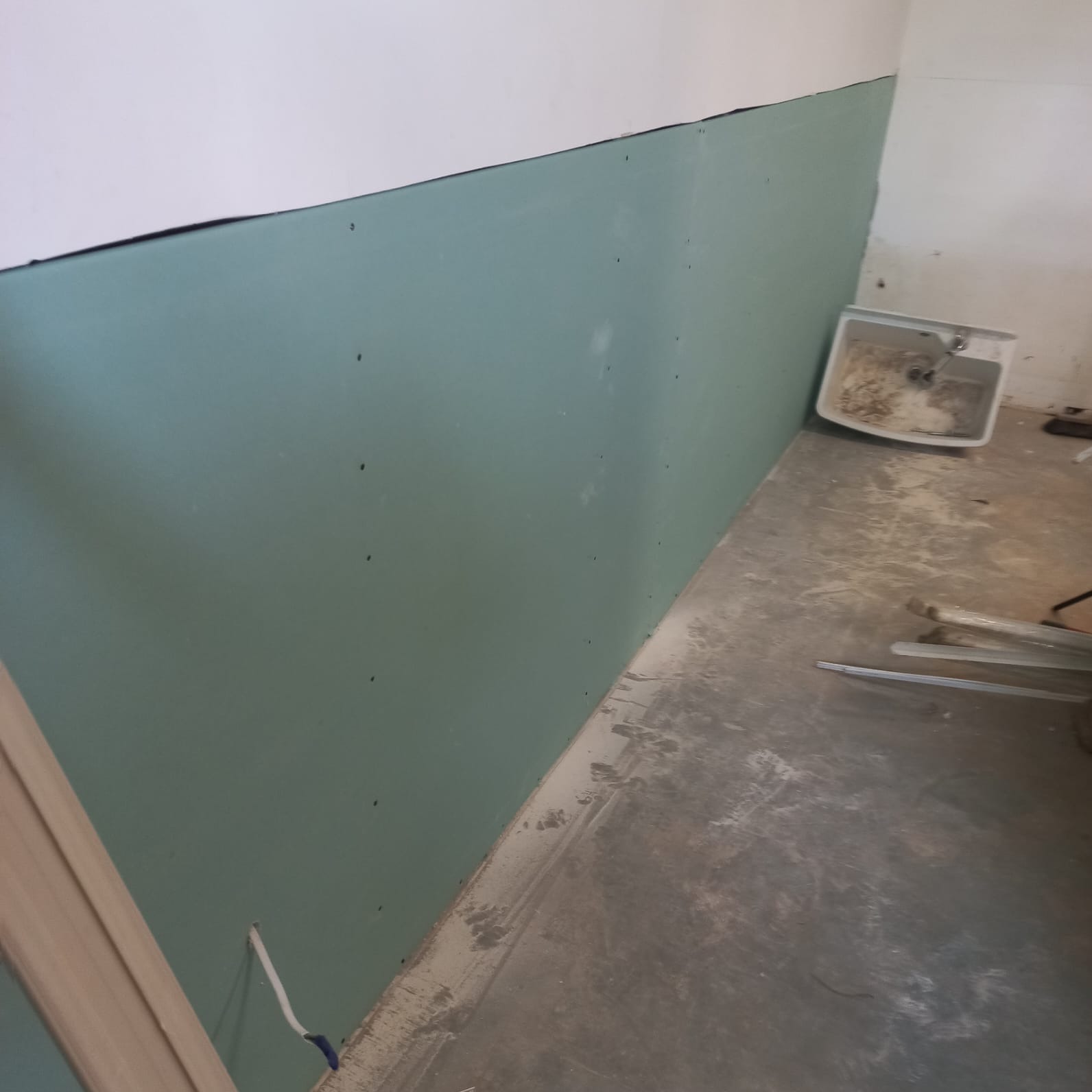 Ściana z płyt gipsowo-kartonowych, z widocznymi śrubami, częściowo pomalowana na biało, z brudną umywalką w rogu i zabrudzoną podłogą w trakcie prac wykończeniowych.