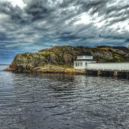 Aneta Panasiuk - Krajobraz morski z kamienistą wyspą, drewnianym pomostem i budynkiem w stylu skandynawskim pod burzowym niebem.