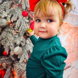 Aneta Panasiuk - Dziewczynka z czerwoną kokardą na głowie, ubrana w zieloną bluzkę, dotyka złotej bombki na tle choinki.