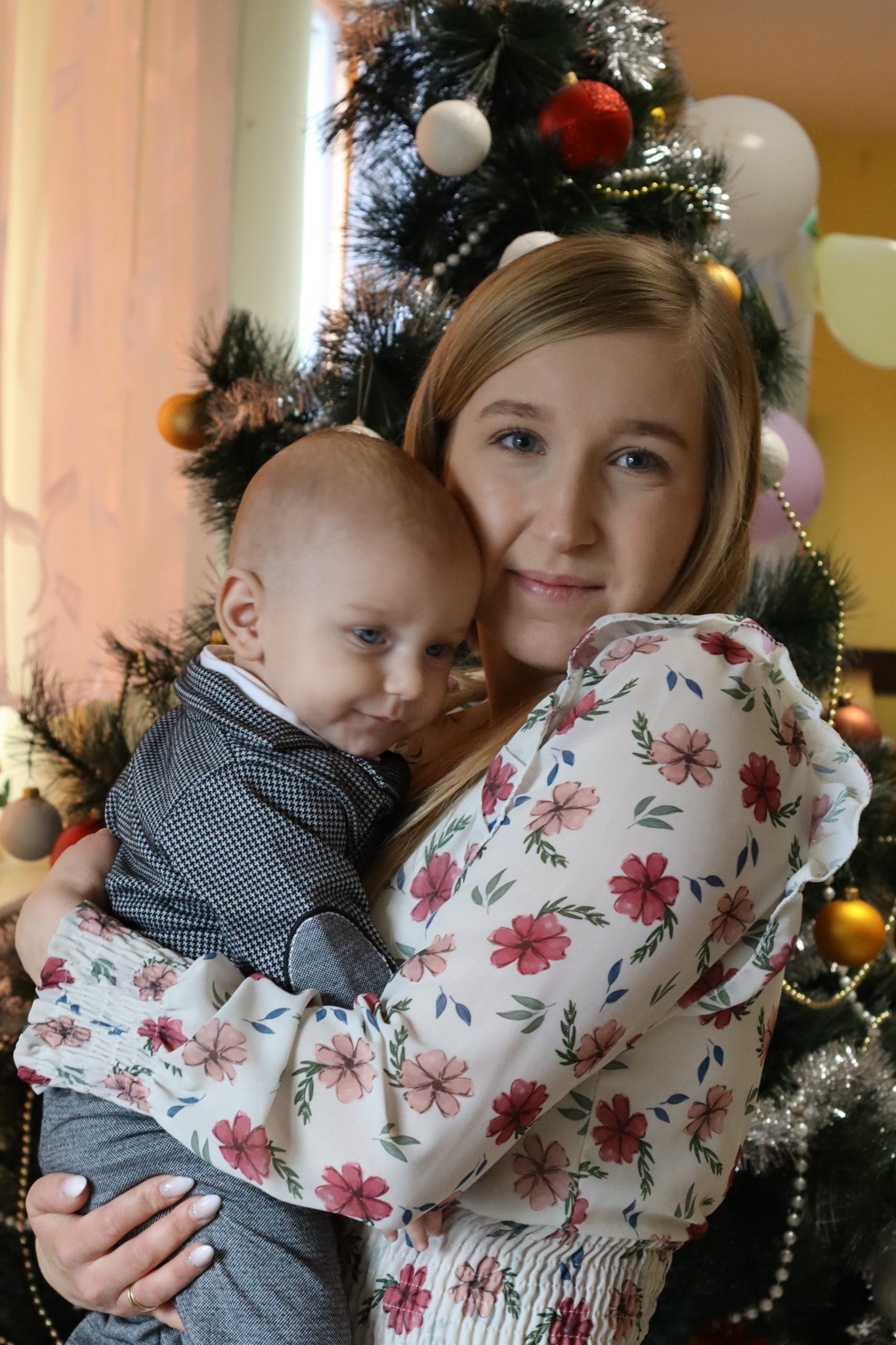 Uśmiechnięta blondynka w kwiecistej bluzce trzyma na rękach niemowlę w eleganckiej marynarce na tle choinki z kolorowymi bombkami i balonami.