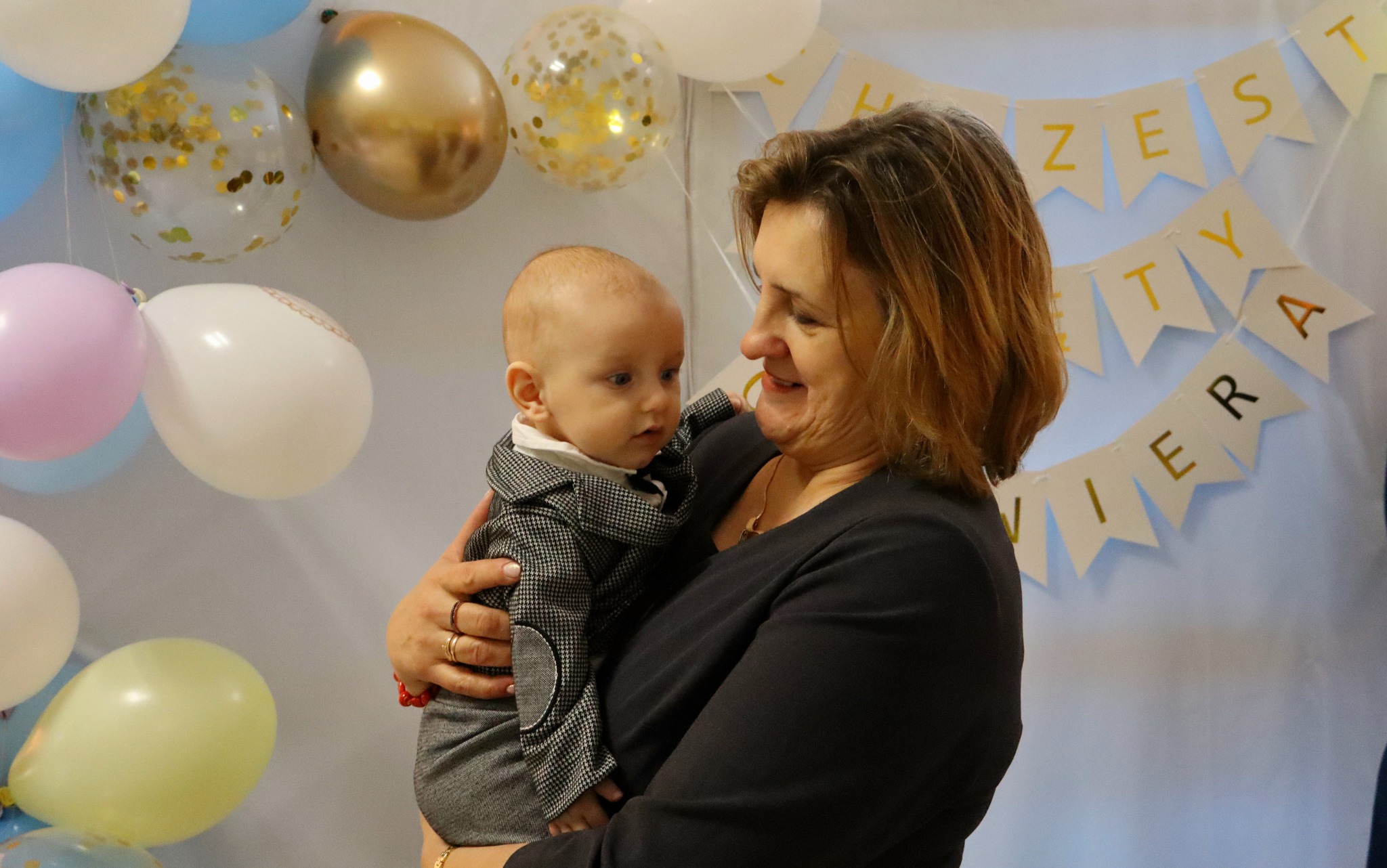 Kobieta trzyma na rękach ubranego w elegancki garnitur niemowlaka na tle balonów i baneru z napisem. Mama patrzy z uśmiechem na dziecko.