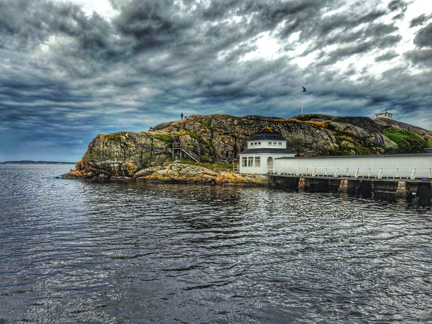 Krajobraz morski z kamienistą wyspą, drewnianym pomostem i budynkiem w stylu skandynawskim pod burzowym niebem.