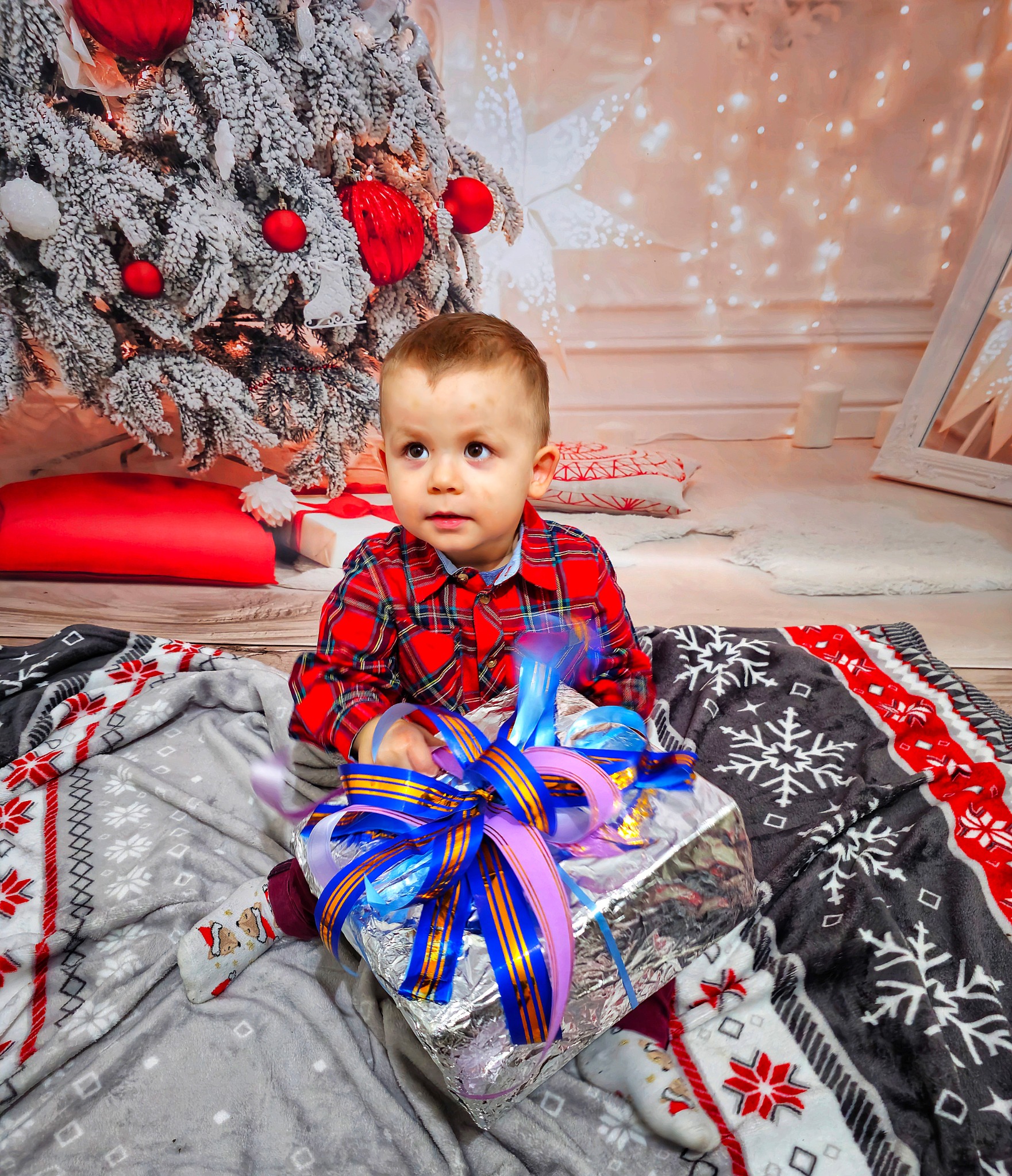 Chłopiec w czerwonej koszuli w kratę siedzi na kocu z prezentem pod choinką, ozdobionym czerwonymi bombkami i sztucznym śniegiem, w tle dekoracja z lampkami.