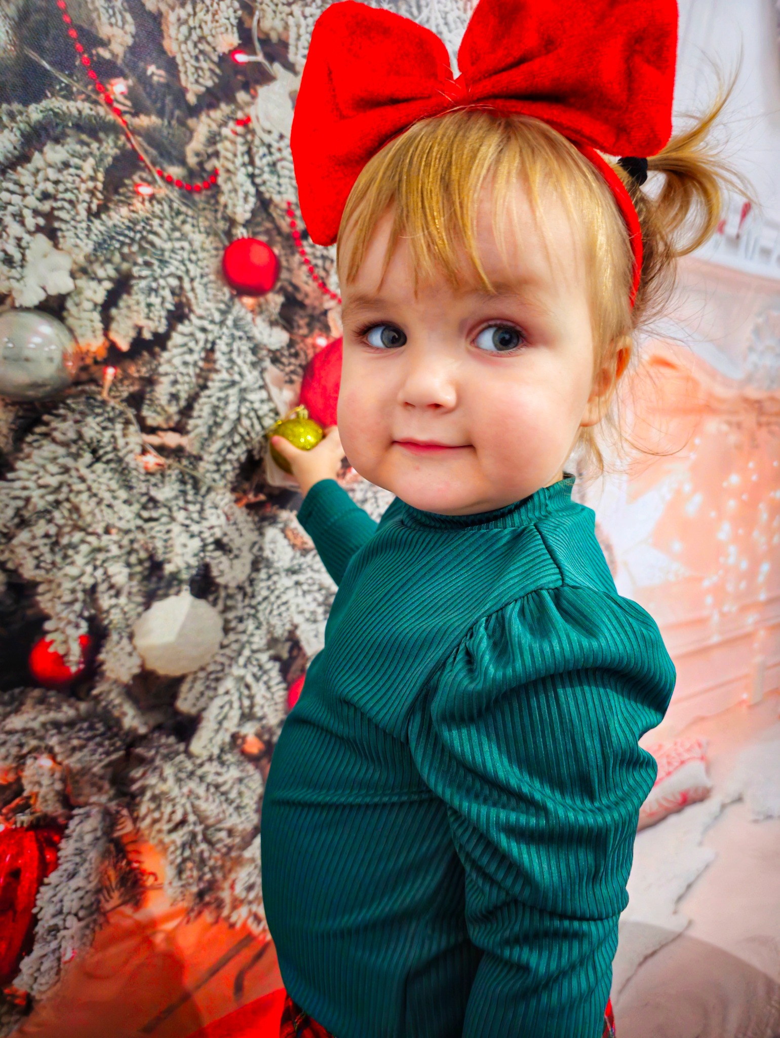 Dziewczynka z czerwoną kokardą na głowie, ubrana w zieloną bluzkę, dotyka złotej bombki na tle choinki.