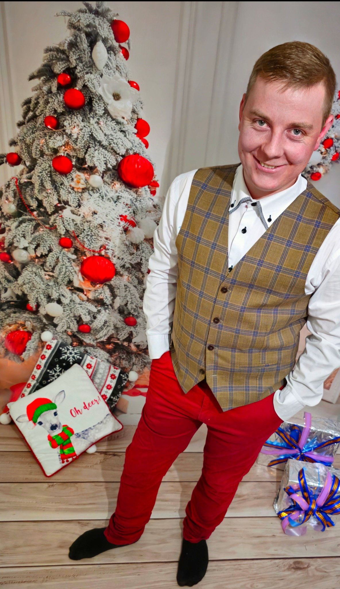 Uśmiechnięty mężczyzna w kamizelce w kratę i czerwonych spodniach pozuje na tle choinki z czerwonymi bombkami i prezentami