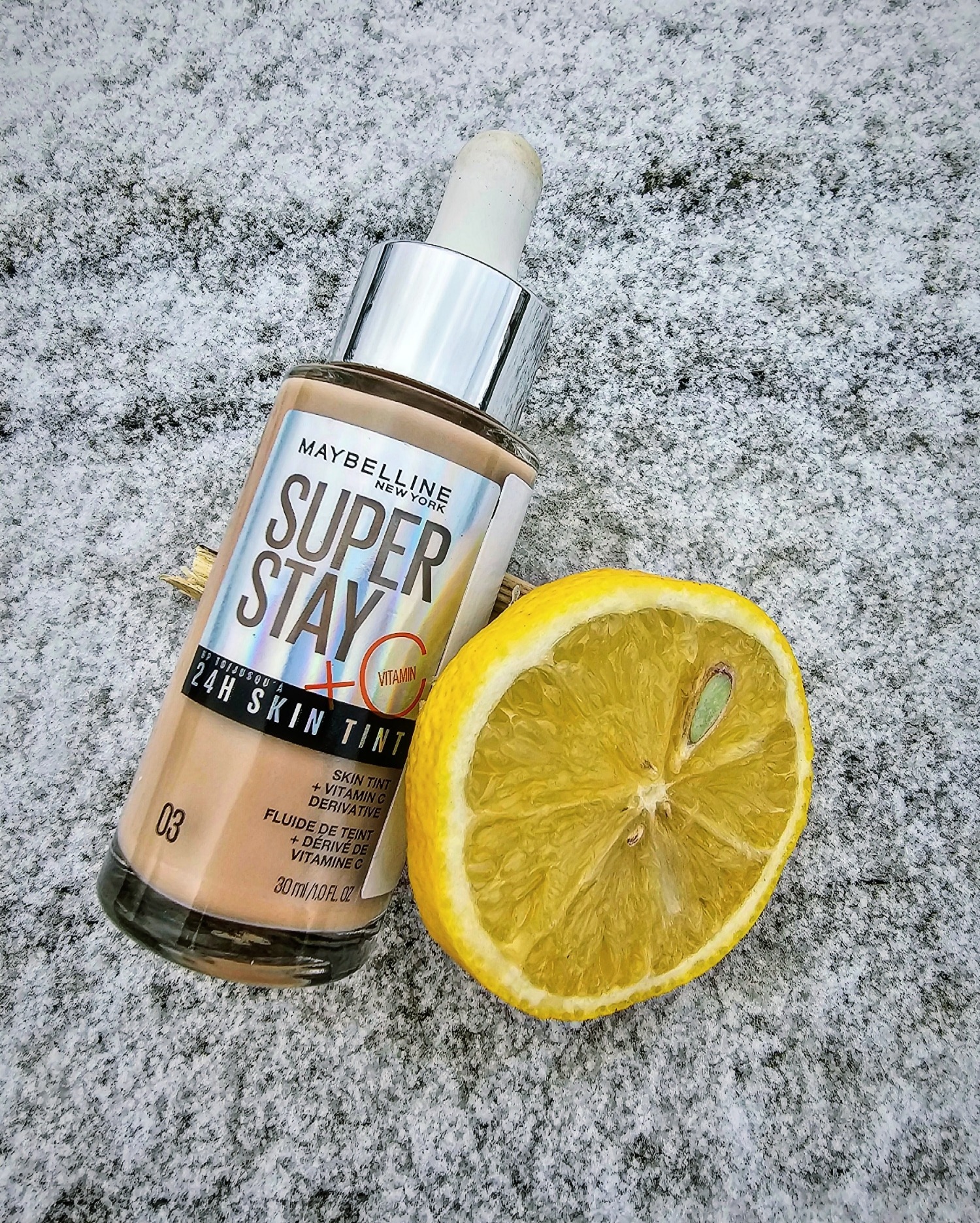 Butelka podkładu Maybelline Super Stay 24H Skin Tint z witaminą C obok połówki cytryny na szaro-białym, ziarnistym tle.
