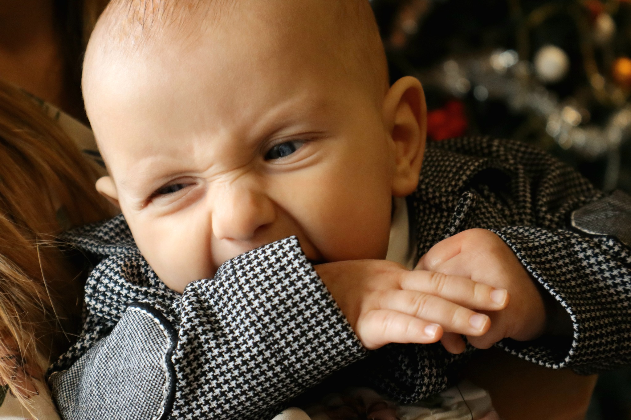 Niemowlę w czarno-białej marynarce w kratkę, z grymasem na twarzy i zaciśniętą dłonią w ustach, w tle rozmazana choinka.