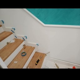 Bartosz Dąbrowski - Montaż drewnianych paneli schodowych: widoczne przycięte stopnice, rolka taśmy malarskiej, nóż monterski i niebieska taśma zabezpieczająca na białych elementach wykończeniowych, ściana pomalowana...