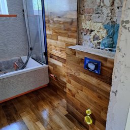 Bartosz Dąbrowski - Montaż drewnopodobnych paneli ściennych w łazience w trakcie remontu; widoczne niedokończone ściany, zamontowana wanna z parawanem i wystające instalacje.