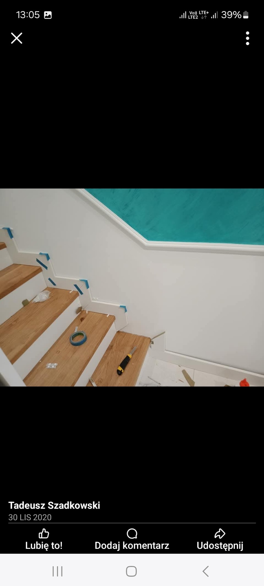 Montaż drewnianych paneli schodowych: widoczne przycięte stopnice, rolka taśmy malarskiej, nóż monterski i niebieska taśma zabezpieczająca na białych elementach wykończeniowych, ściana pomalowana...