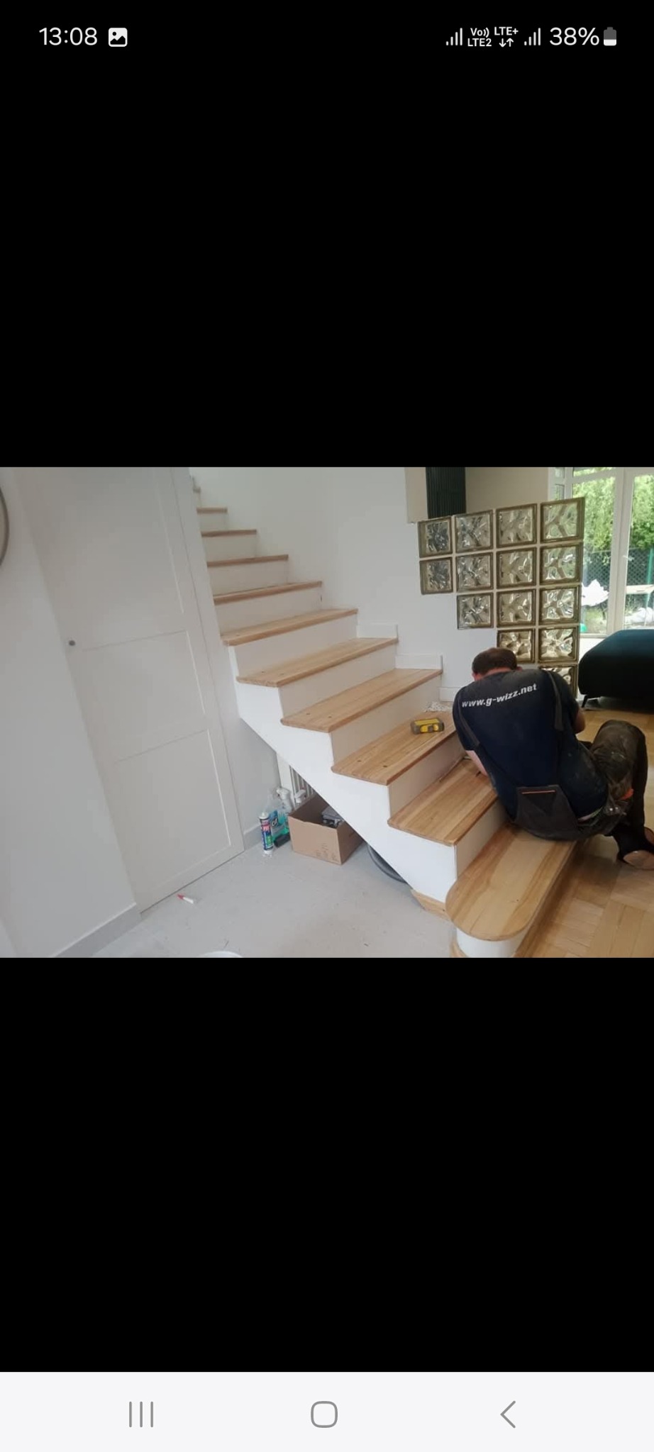 Pracownik w ciemnej koszulce z logo firmy szlifuje drewniane stopnie schodów prowadzących na górę, w tle ściana z luksferami i drzwi balkonowe.