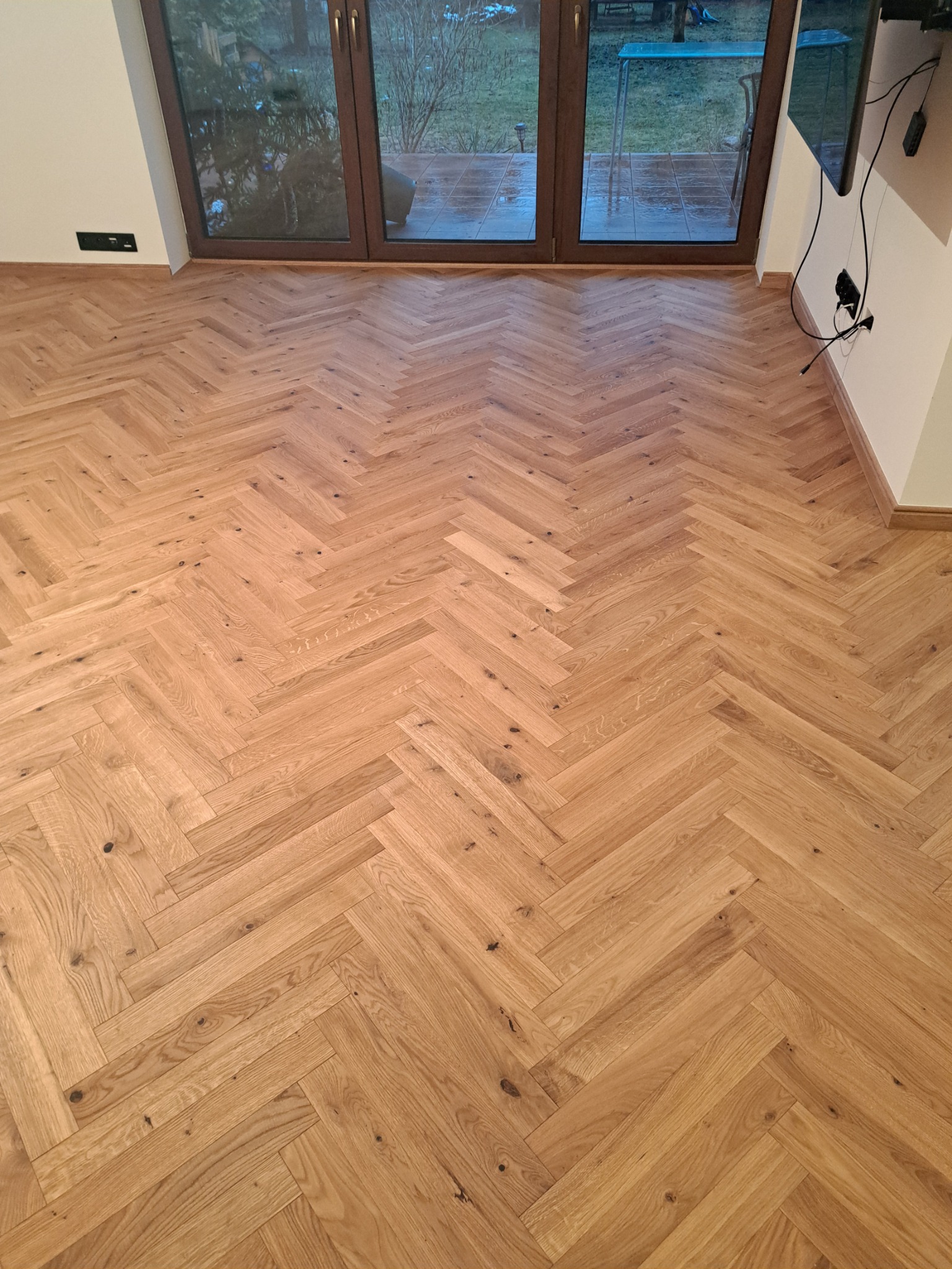 Ułożona w jodełkę, jasna drewniana podłoga parkietowa, widok na okno balkonowe i fragment ściany z gniazdkami elektrycznymi.