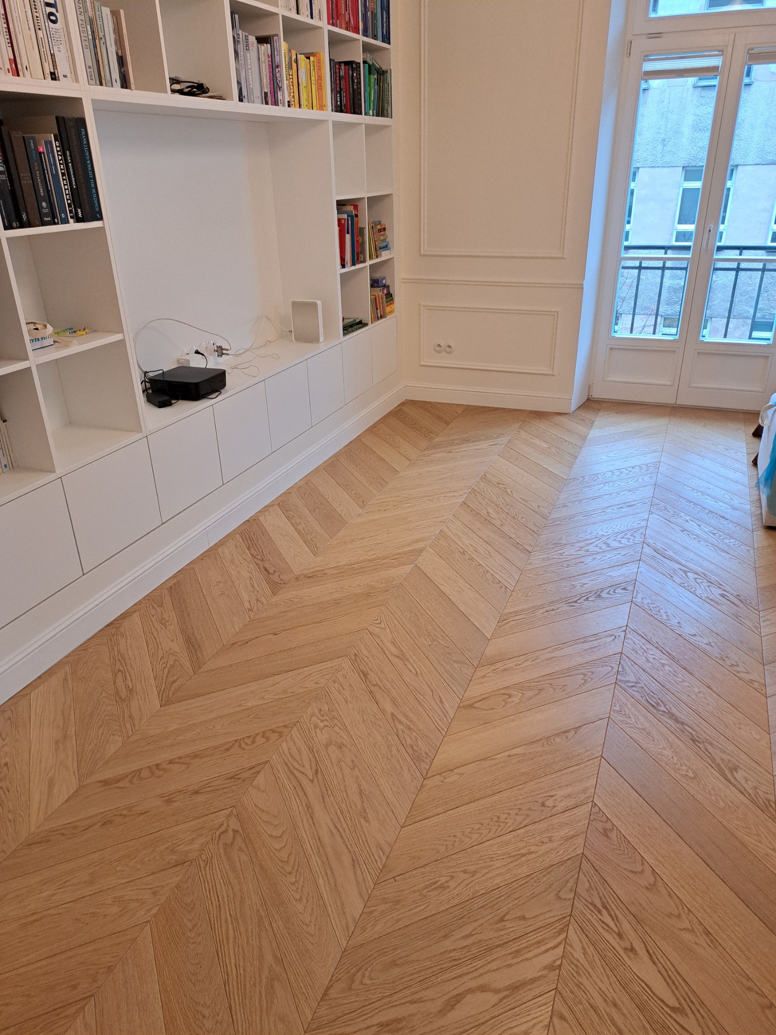 Jasny parkiet ułożony we wzór jodełki francuskiej w przestronnym salonie z białymi meblami w zabudowie i dużym oknem balkonowym.