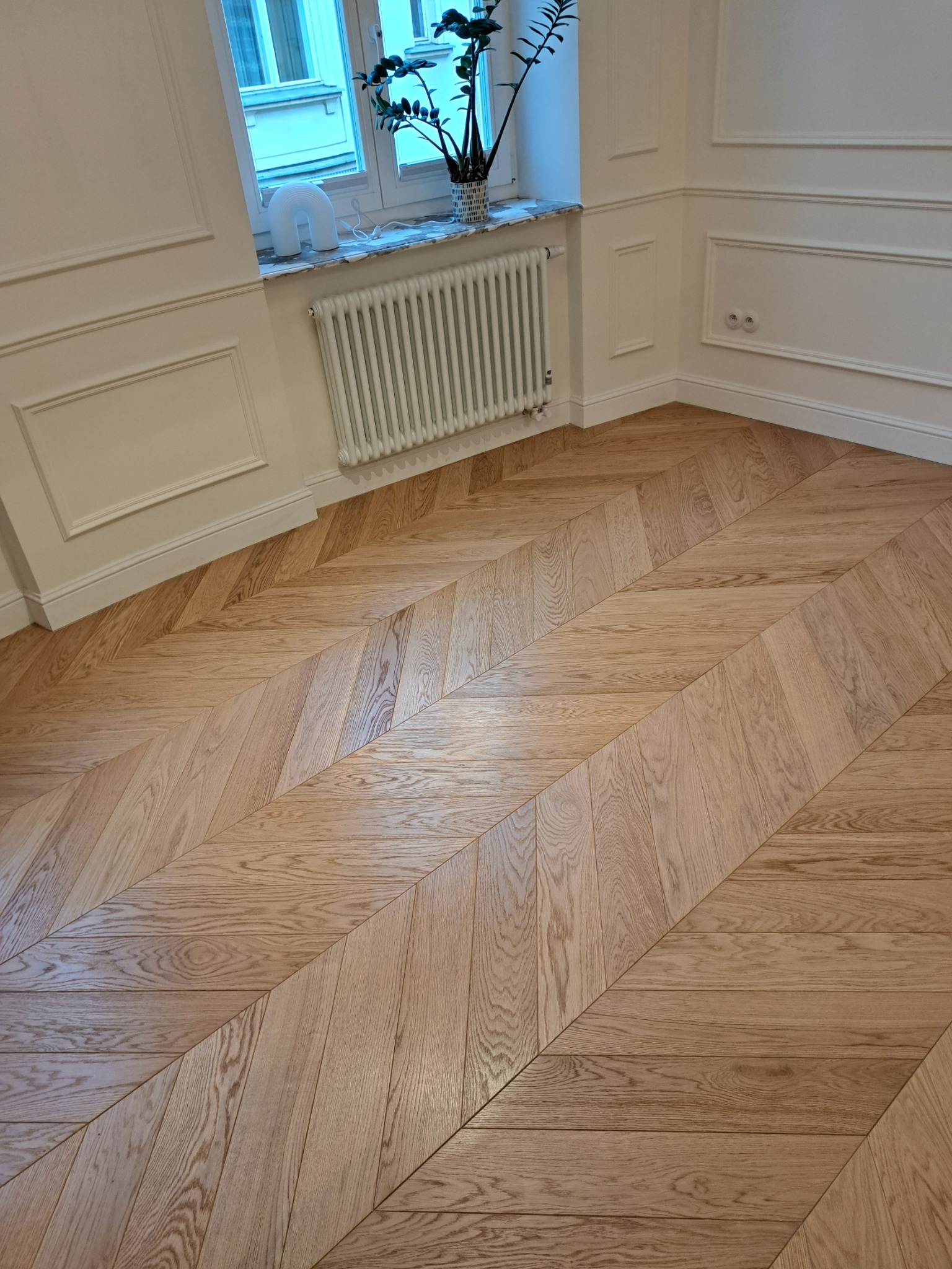 Jasny parkiet ułożony we wzór jodełki w rogu pokoju z białymi ścianami z dekoracyjnymi listwami i grzejnikiem pod oknem.