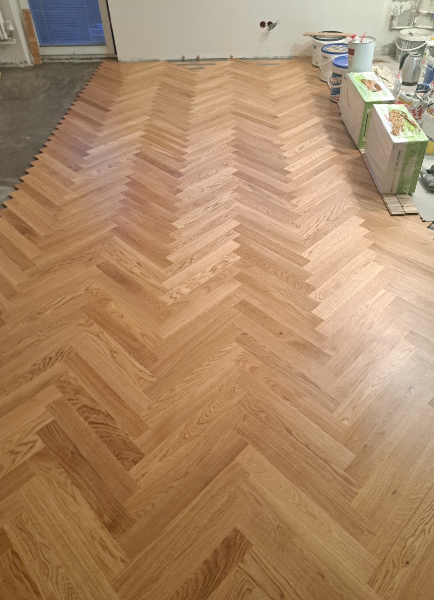 Układanie parkietu w jodełkę, widoczne puste przestrzenie i niedokończone krawędzie, w tle puszki z farbą i narzędzia malarskie.