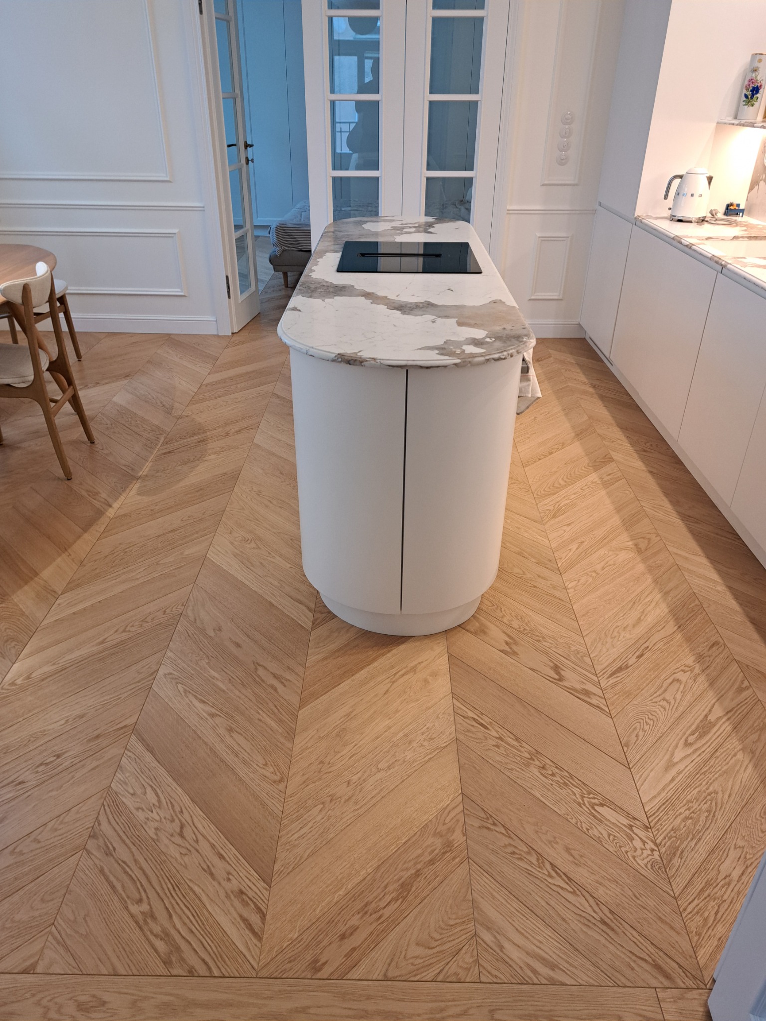 Jasny parkiet ułożony we wzór jodełki francuskiej w nowoczesnej kuchni z białą wyspą i zabudową.